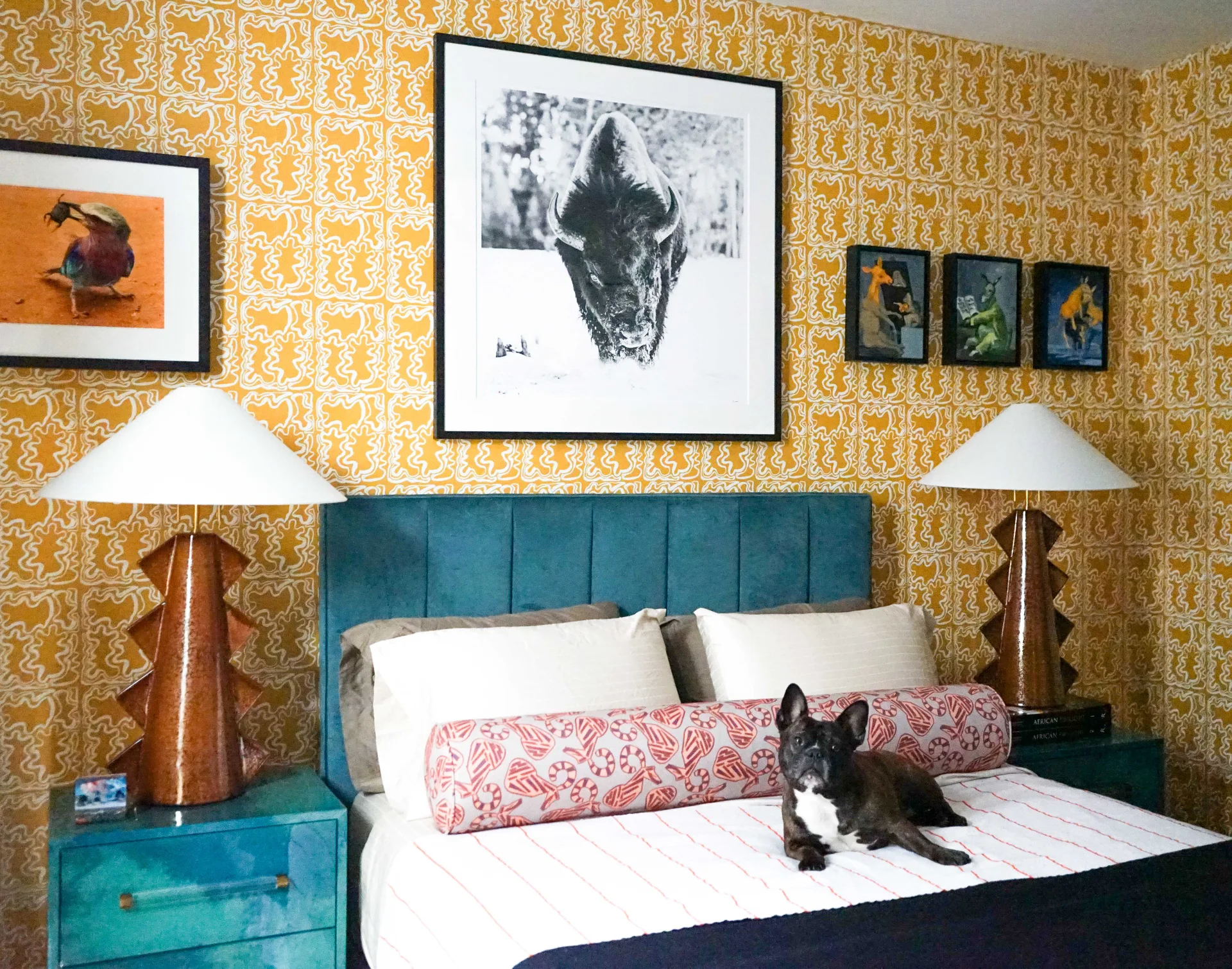 An eclectic bedroom features yellow patterned wallpaper, a teal upholstered headboard, a pair of sculptural brown lamps, and a black French bulldog on the bed.