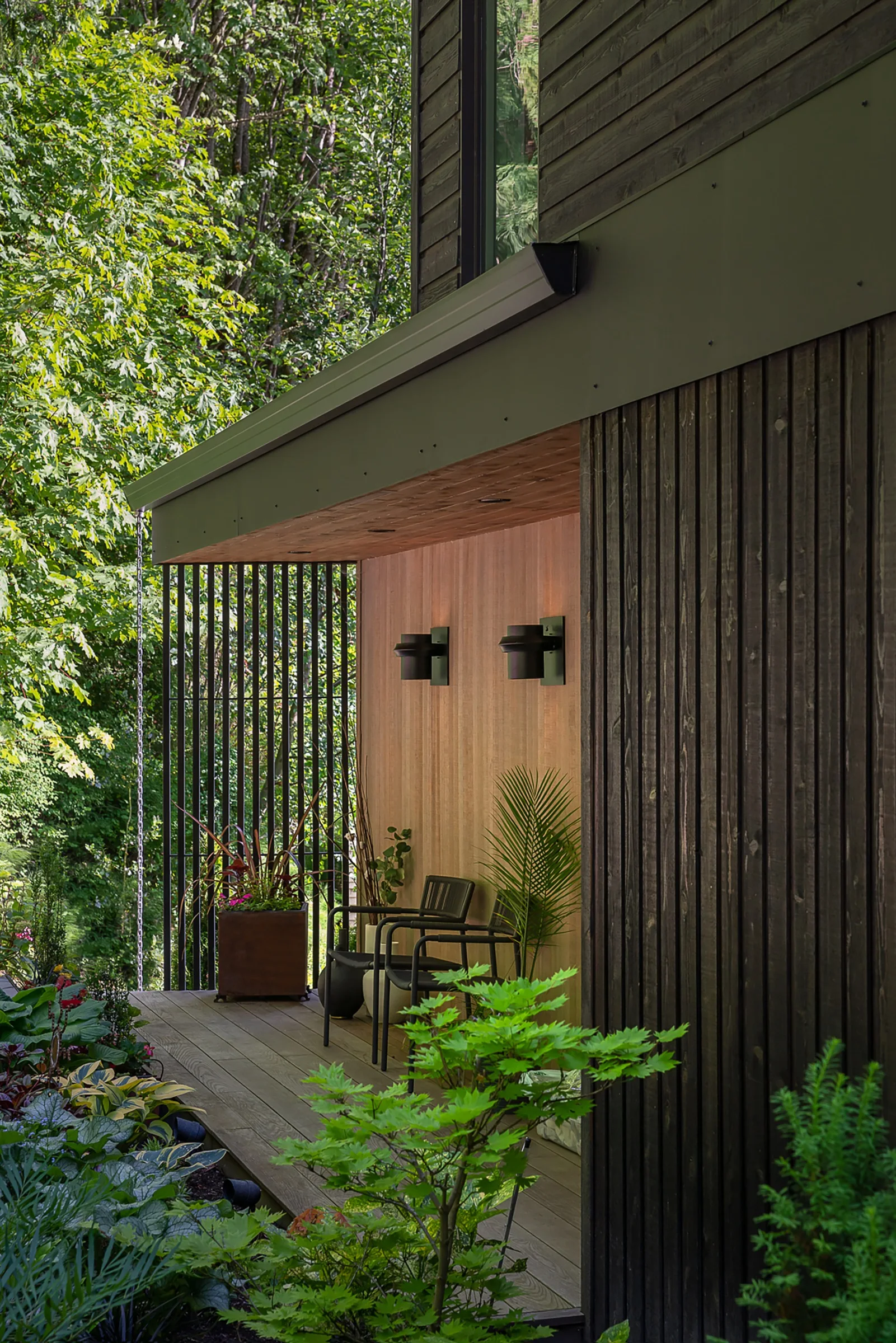 A covered wooden porch features dark vertical wood siding, two black metal armchairs beneath warm wall sconces, and lush green plants.