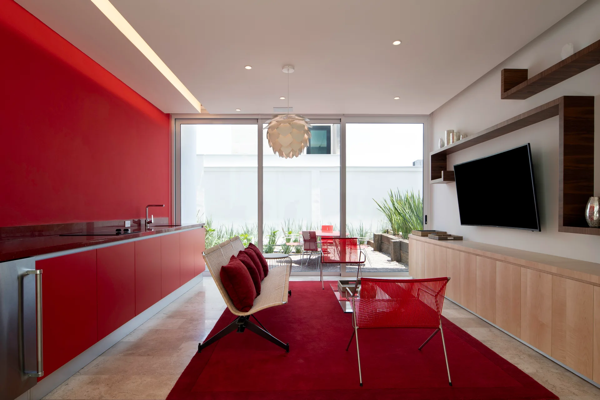 A modern living room features a vibrant red kitchen cabinet wall, a coordinating red area rug, light wood console, and sliding glass doors to a patio.