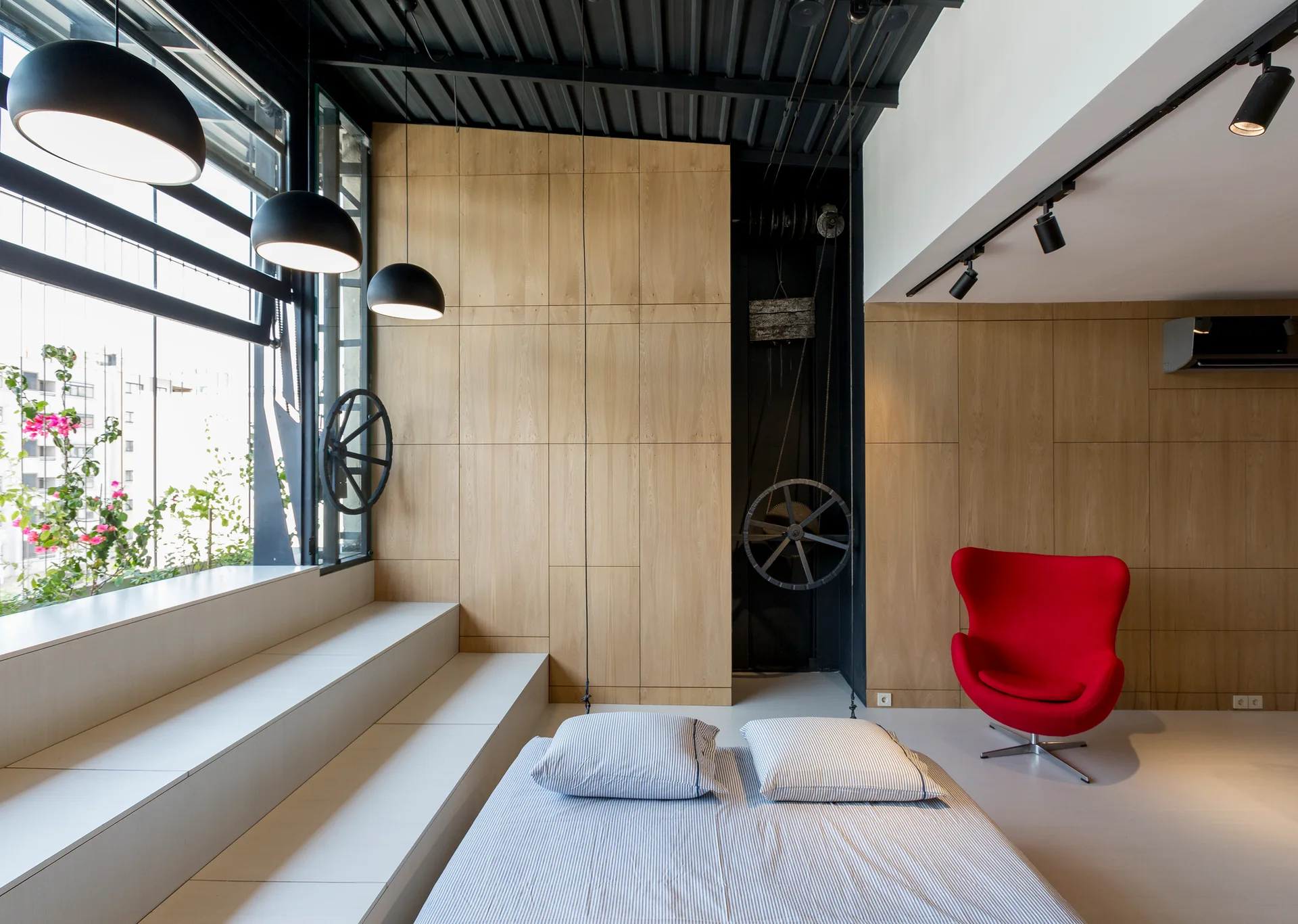 A modern bedroom featuring a bed with striped bedding next to a stepped white platform, pale wood wall panels, and a red armchair.