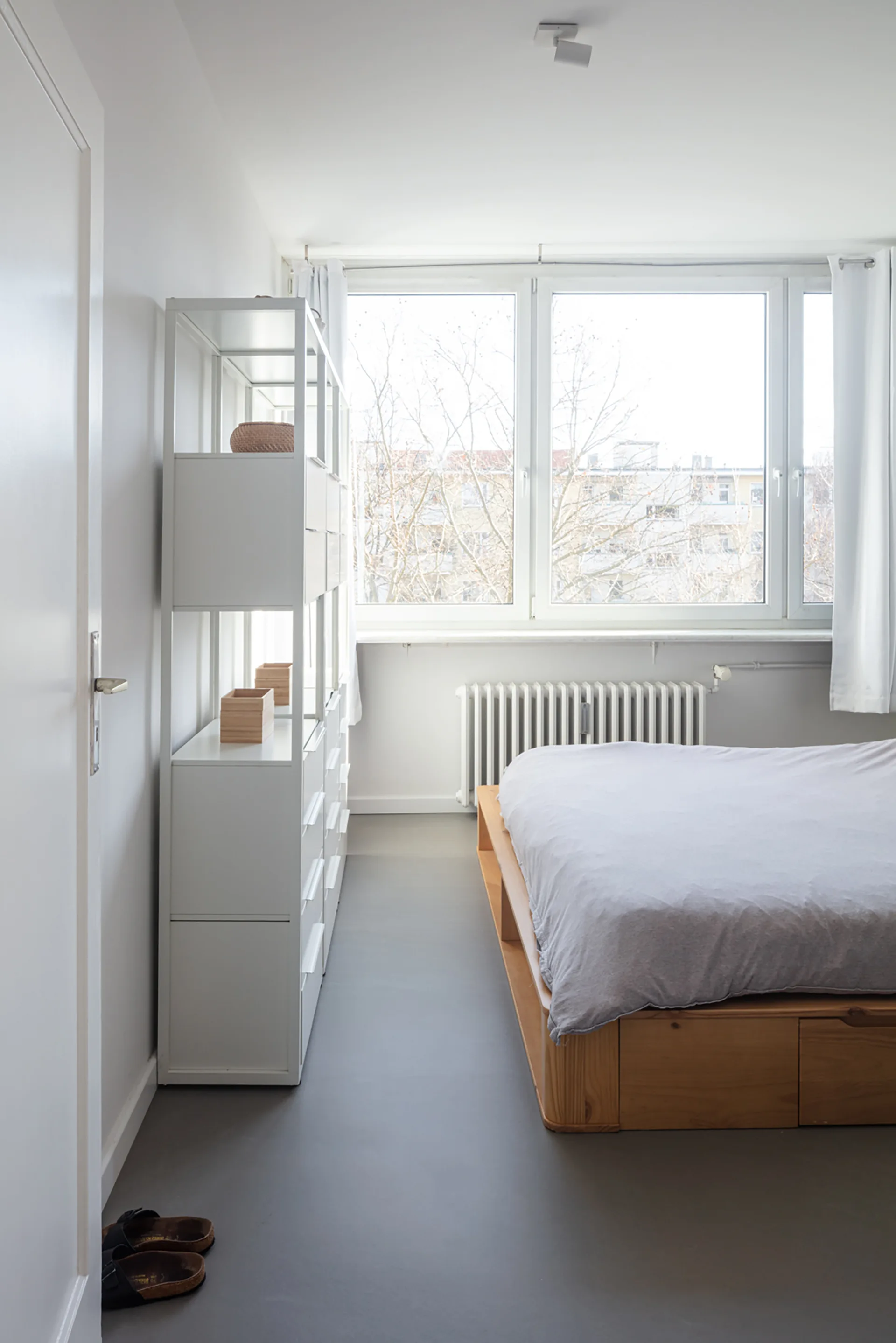 A minimalist bedroom featuring a wooden platform bed with grey bedding, a tall white shelving unit, and a large window.