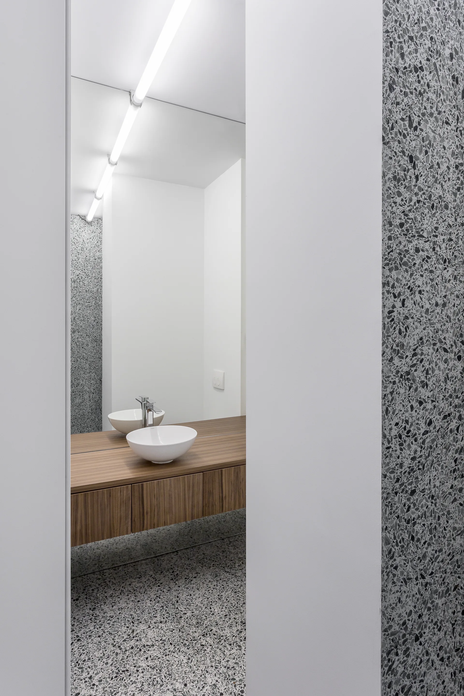 A minimalist bathroom features a floating wood-grain vanity, a white vessel sink, a linear light fixture, and speckled grey terrazzo flooring.
