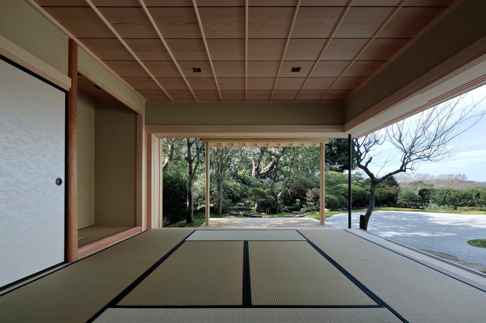 A traditional Japanese room features woven tatami mat flooring with black borders, a wood grid ceiling, and a wide opening overlooking a green garden.