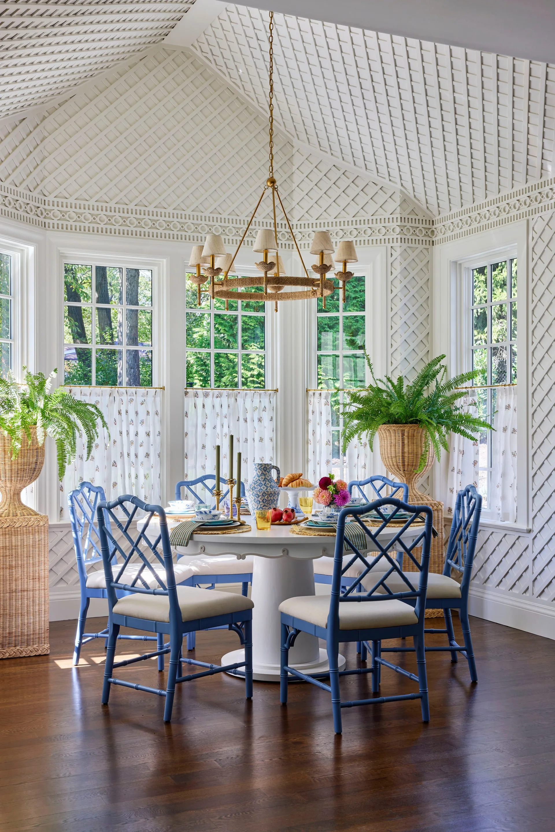 A sunlit dining room features a white pedestal table and blue bamboo-style chairs surrounded by white lattice-covered walls and a vaulted ceiling.
