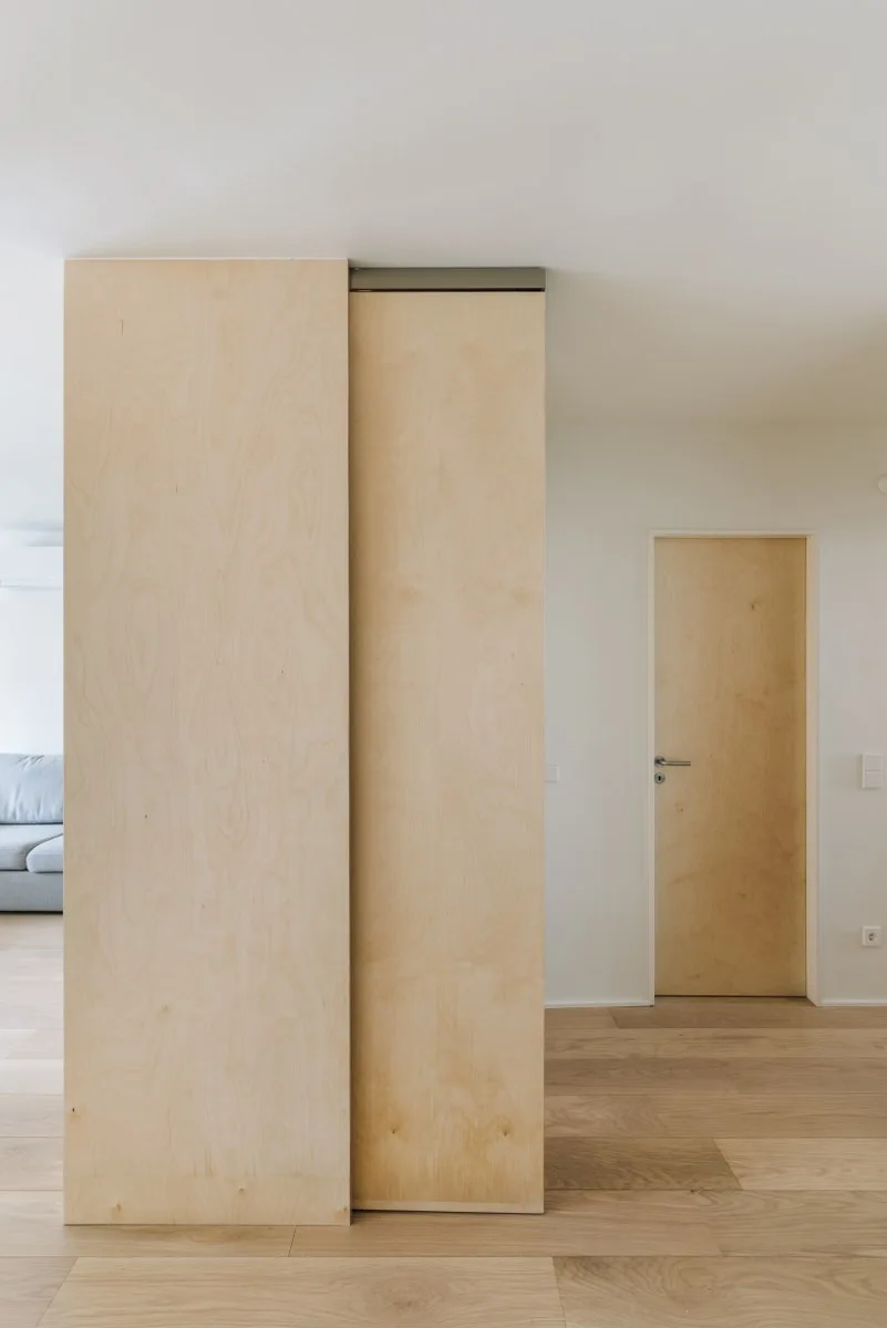 A minimalist interior featuring a large light plywood room divider and a matching wooden door set against white walls and light wood floors.