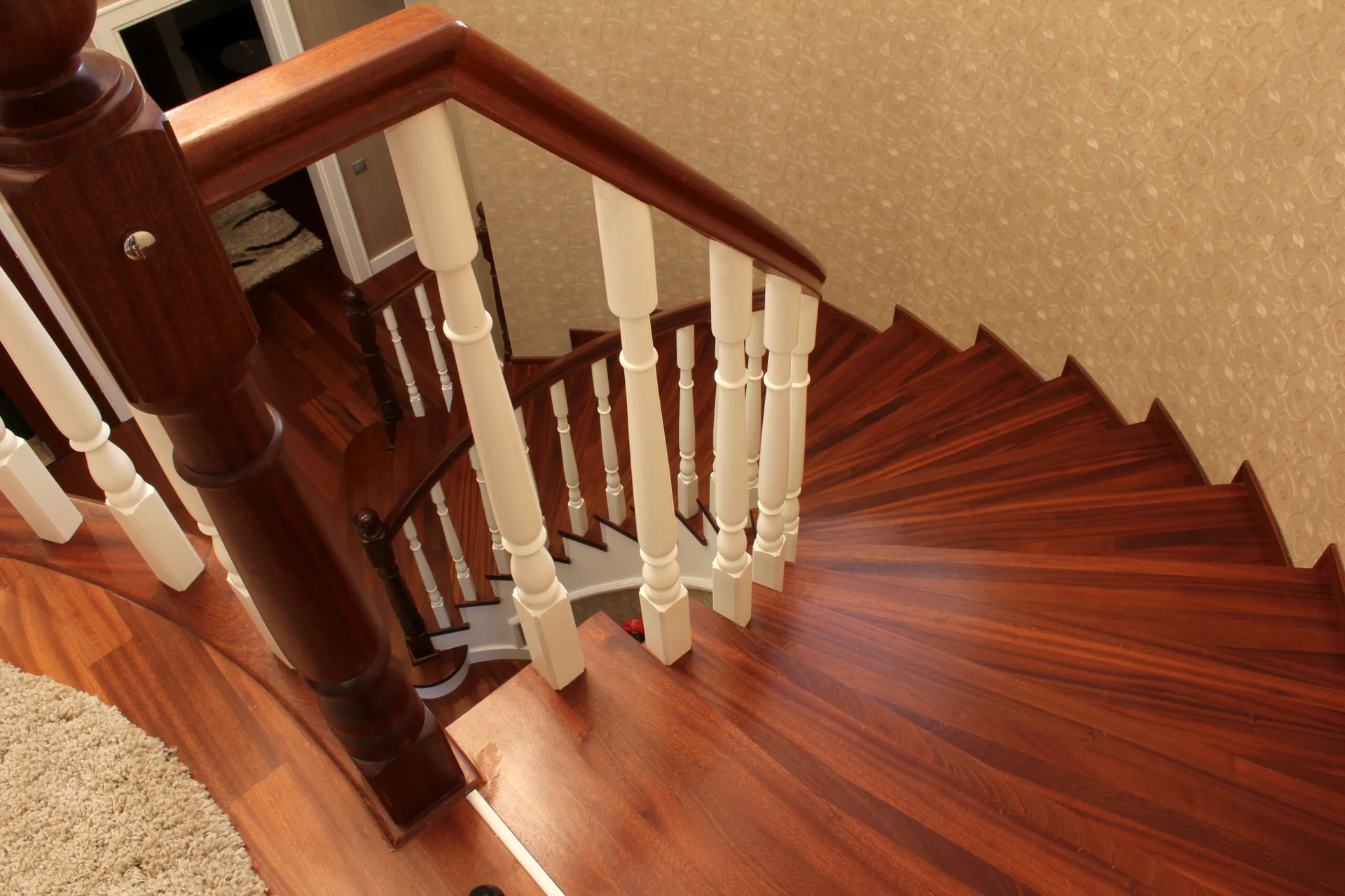 A top-down view of a curved wooden staircase with dark mahogany treads, a matching handrail, and white painted balusters against a patterned beige wall.