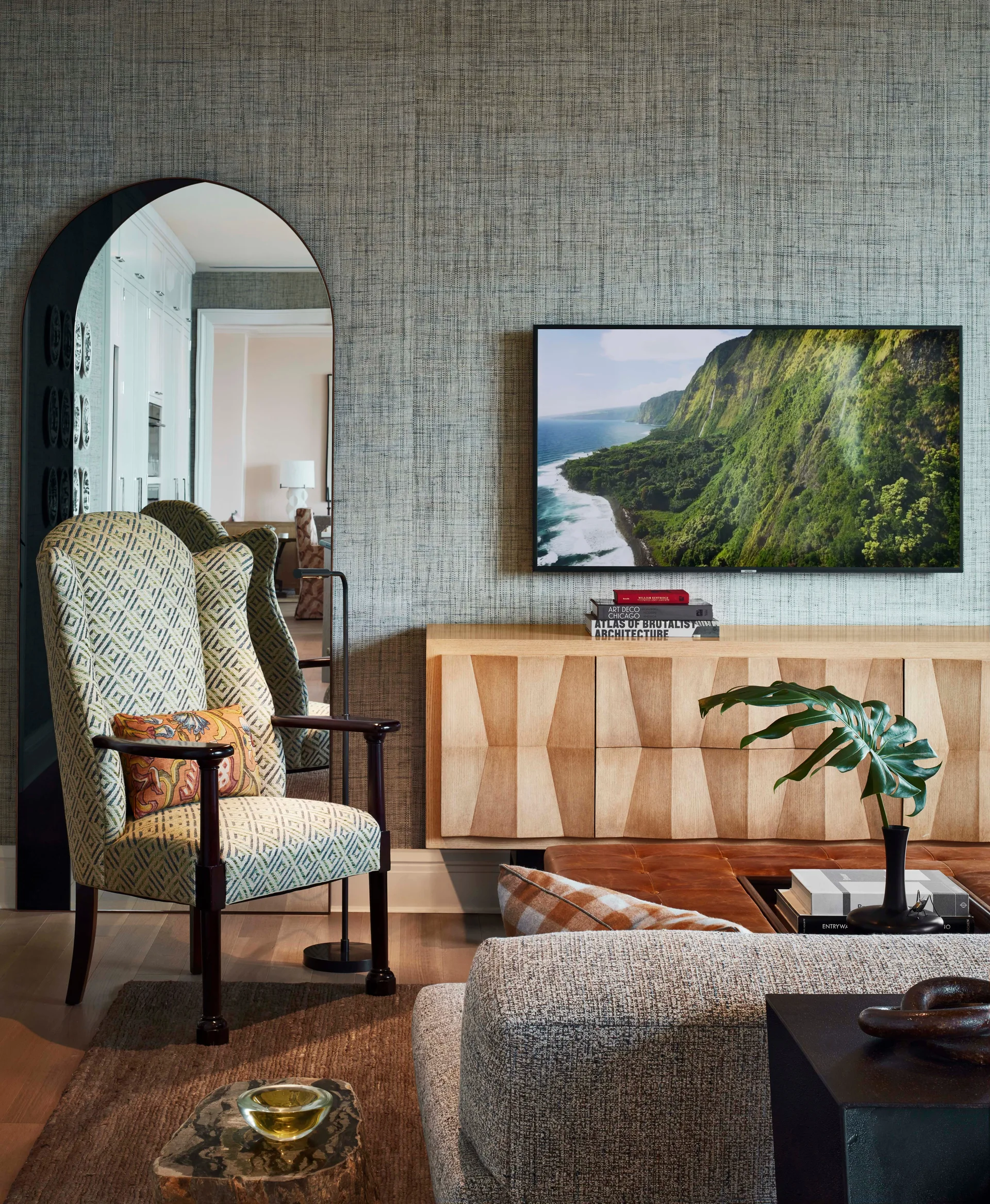 A media room features a wall-mounted TV over a light wood geometric console, an arched floor mirror, and a patterned wingback chair.