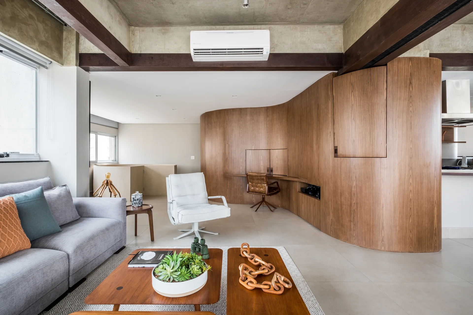 A modern living room features a grey sofa, a curved wood paneled wall with a built-in desk, and an exposed concrete ceiling with dark beams.