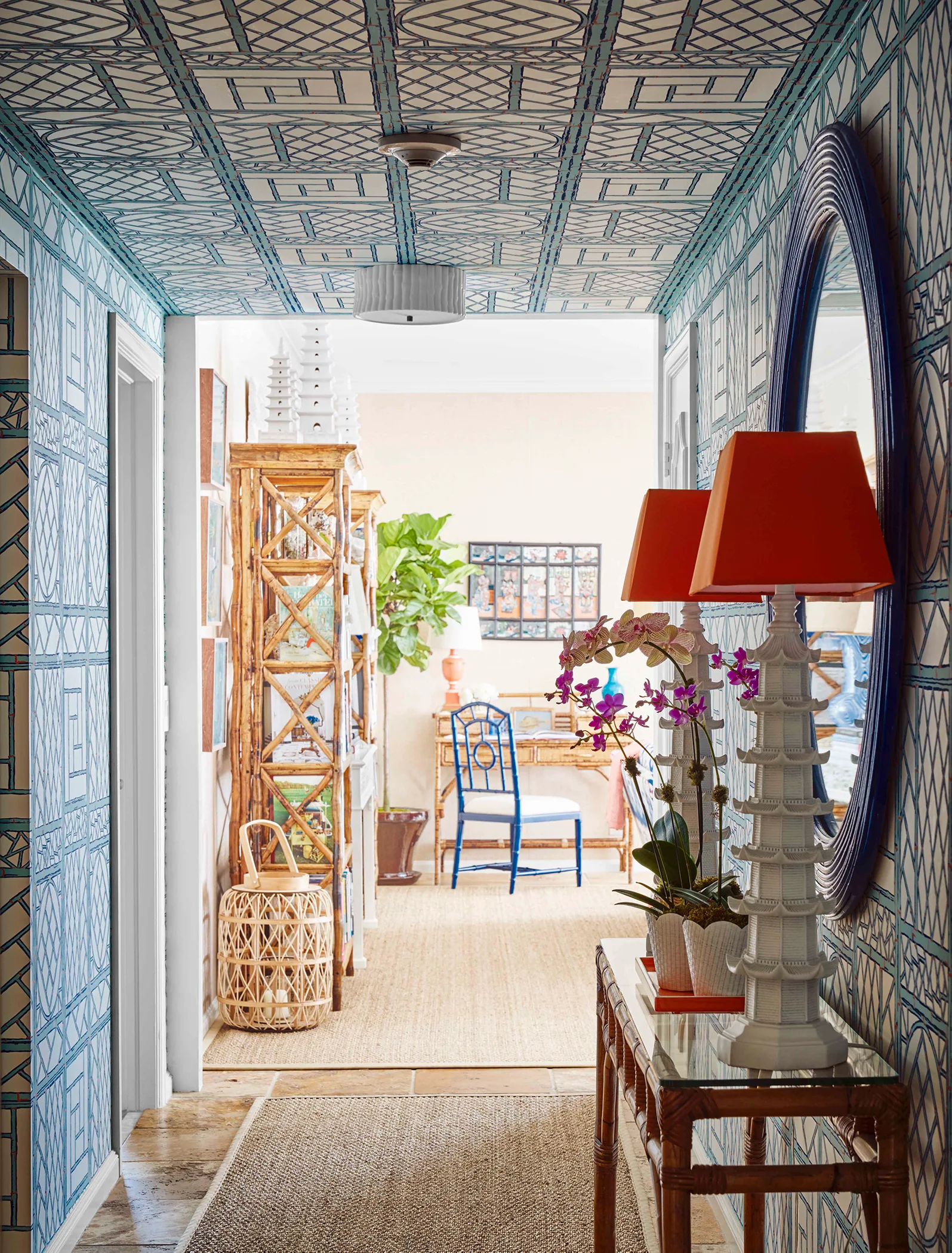 A hallway lined with blue and white trellis wallpaper on the walls and ceiling features a bamboo console table with two orange-shaded pagoda lamps.
