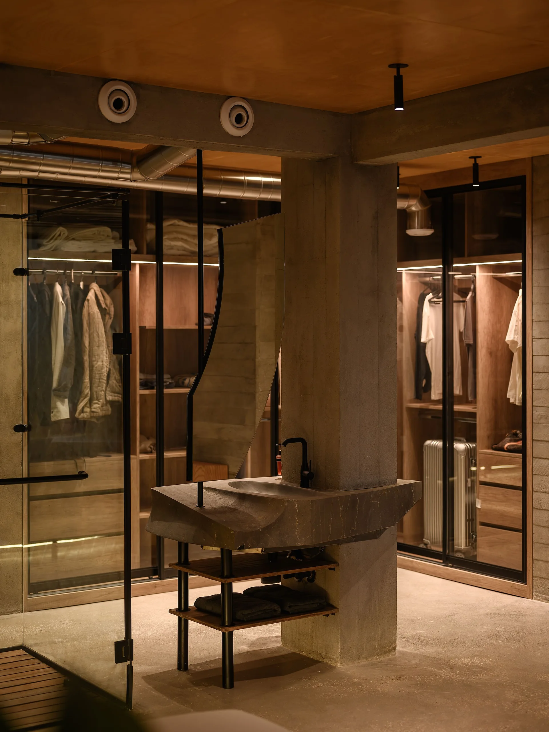 A modern dressing room featuring a gray stone vanity mounted to a concrete pillar, with lighted wooden wardrobes and a glass partition in the background.