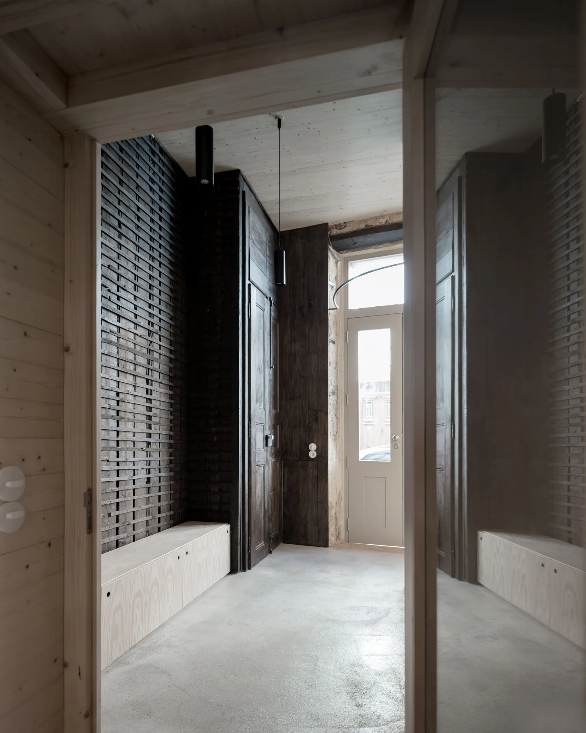 A minimalist entryway features a light wood bench below a dark slatted accent wall, concrete floors, and a glass-paneled entry door.