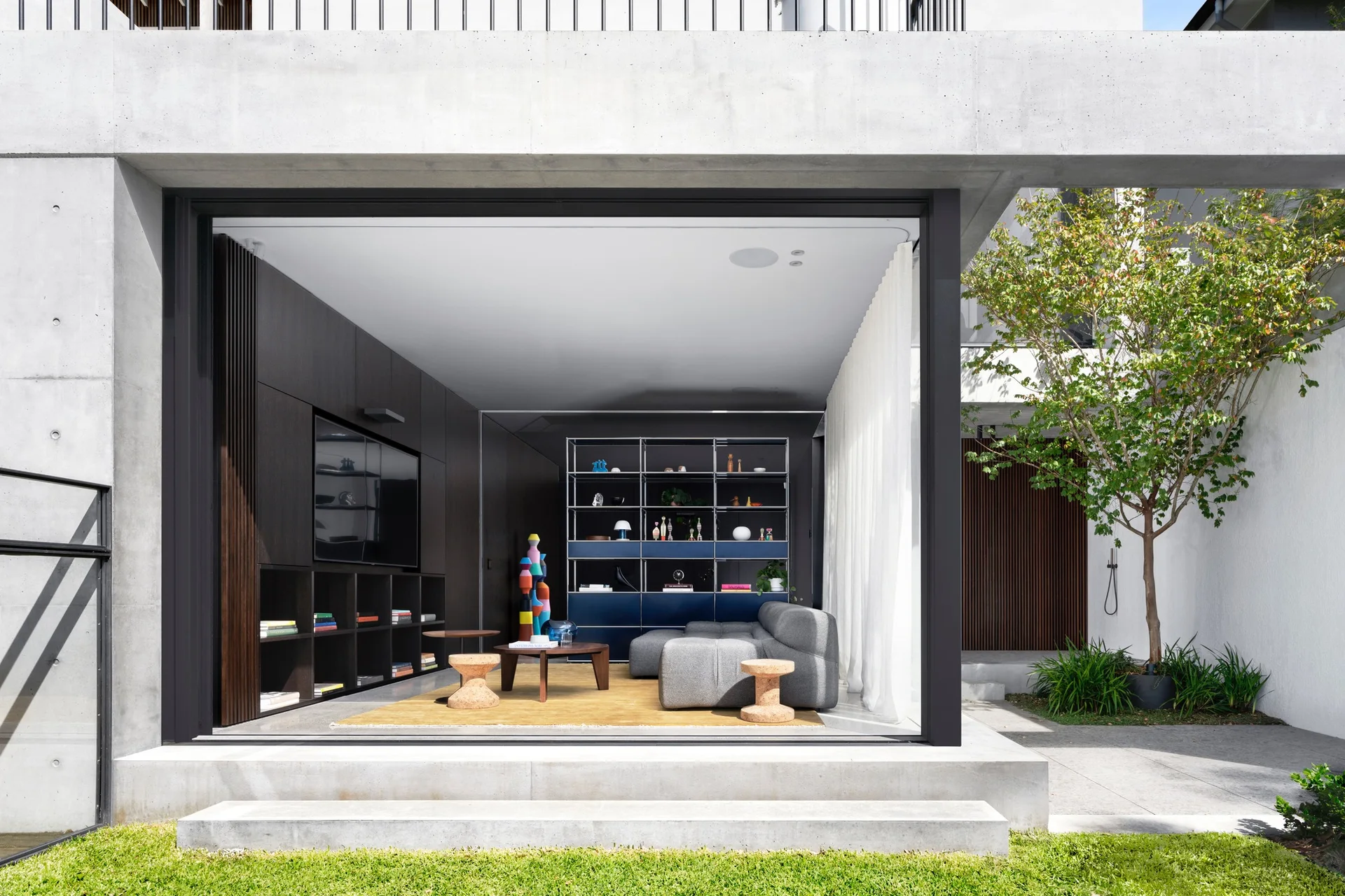 A modern living room with a gray sectional and dark wood media wall opens to a concrete courtyard through a sliding glass door.