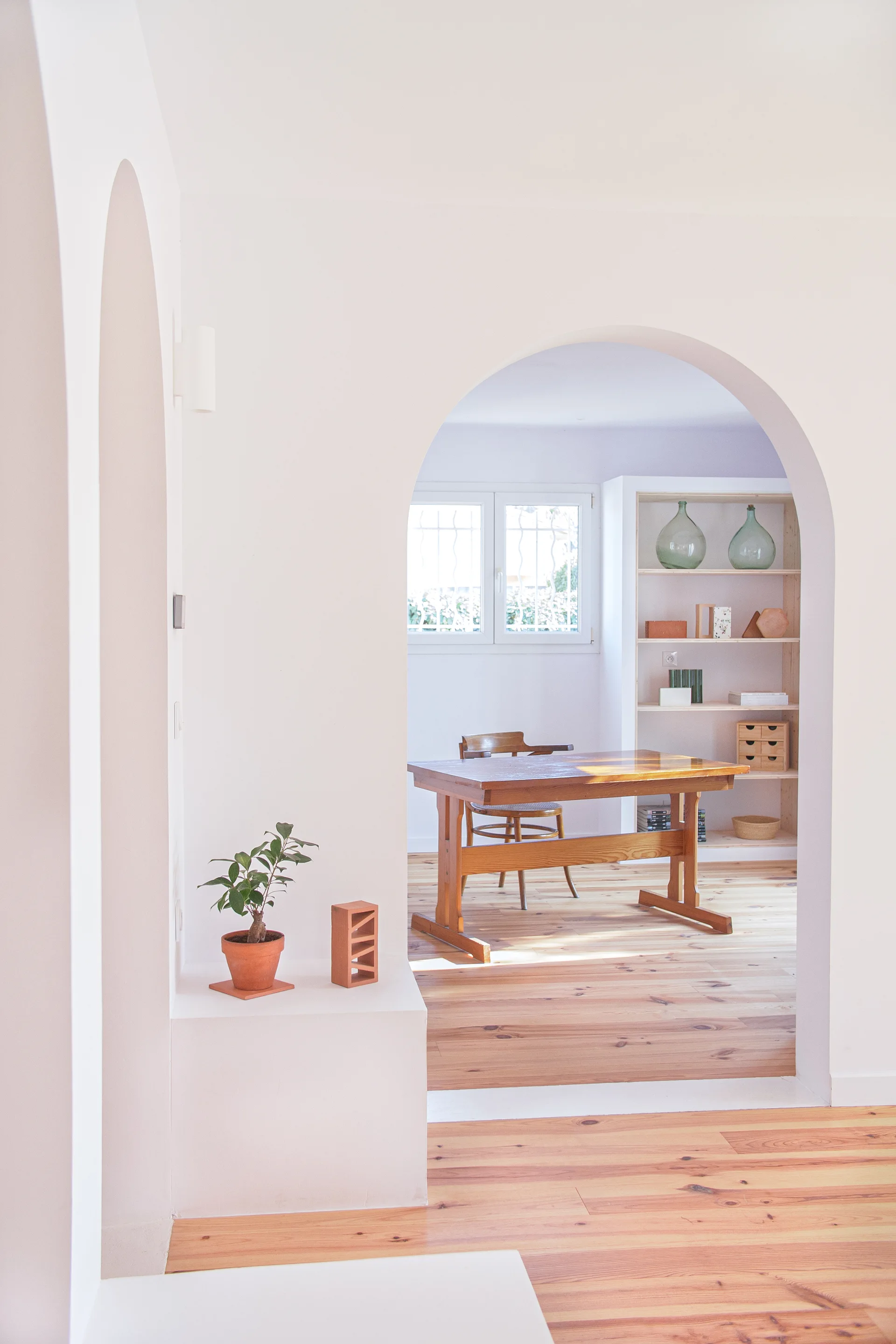 A smooth plaster archway frames a minimalist dining room featuring a wooden table, a matching chair, and light wood plank floors.