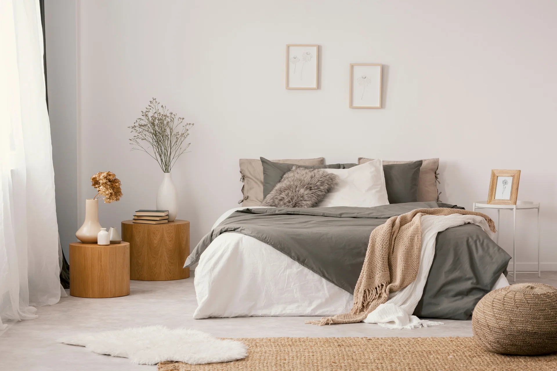 A minimalist bedroom features a bed with grey and white bedding, two cylindrical wood side tables, and a woven rug on the floor.