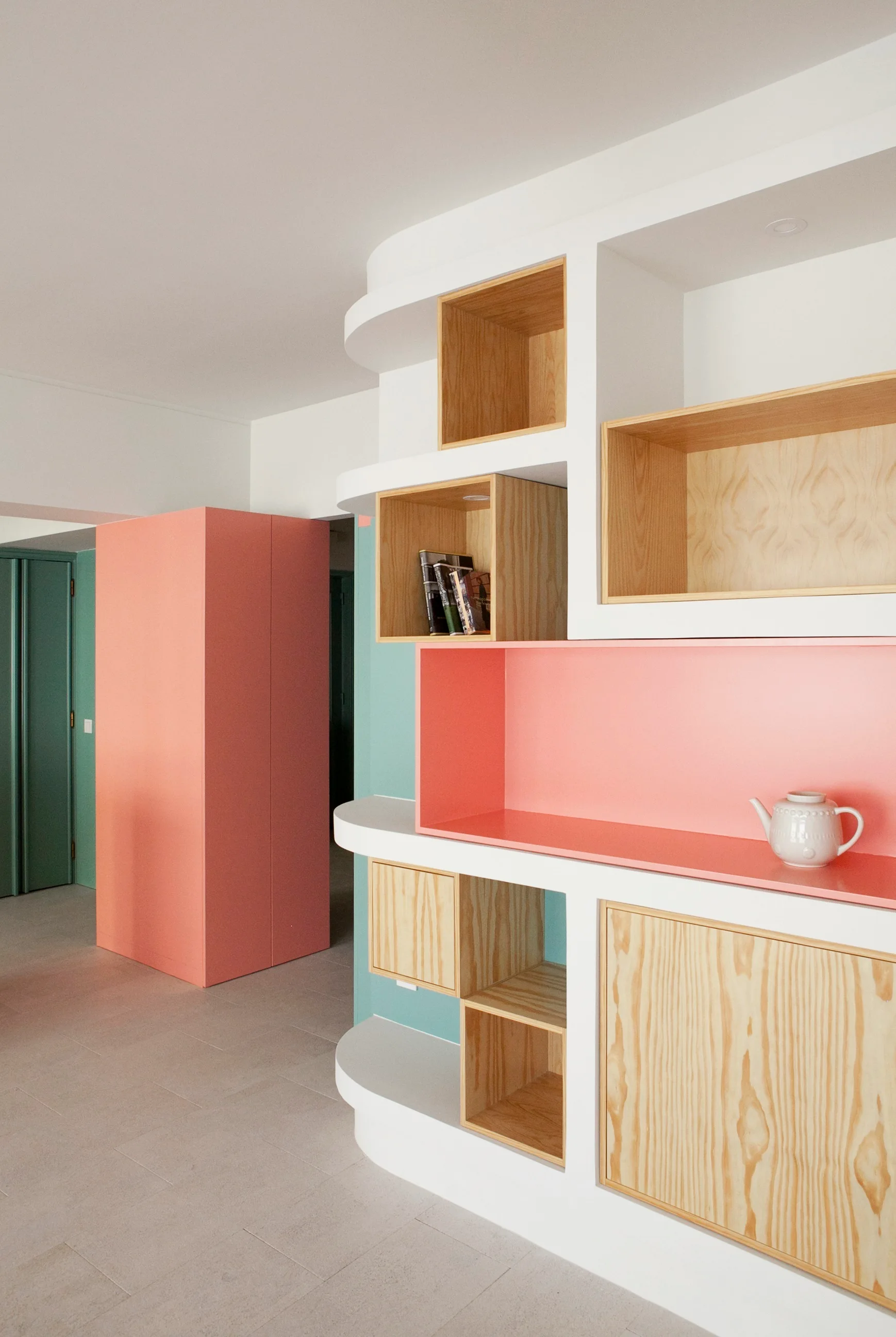 A white curved built-in storage unit features plywood cubbies and a pink shelf next to a matching pink cabinet and teal walls.