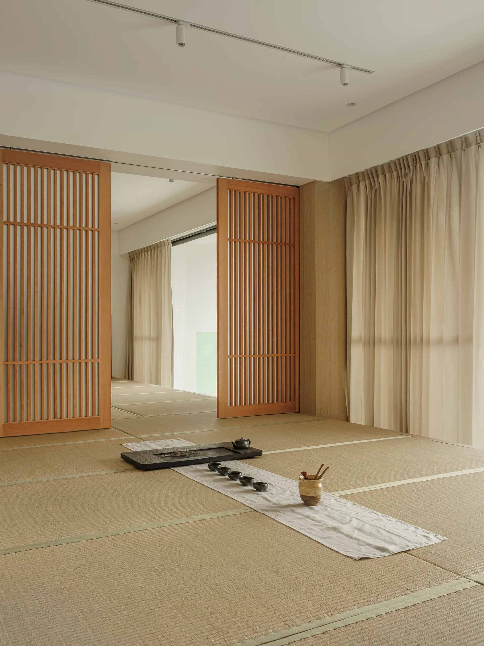 A minimalist tatami room featuring light wood slatted sliding doors, neutral drapes, and a black ceramic tea set arranged on a low floor runner.