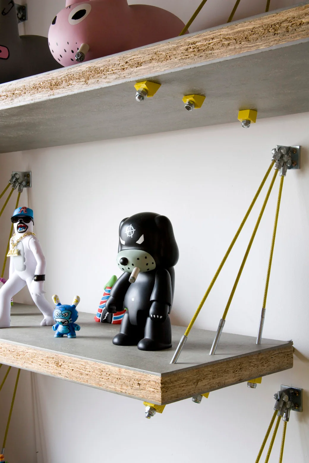Floating grey shelves with exposed wood edges and yellow wire supports hold stylized plastic art figurines against a white wall.