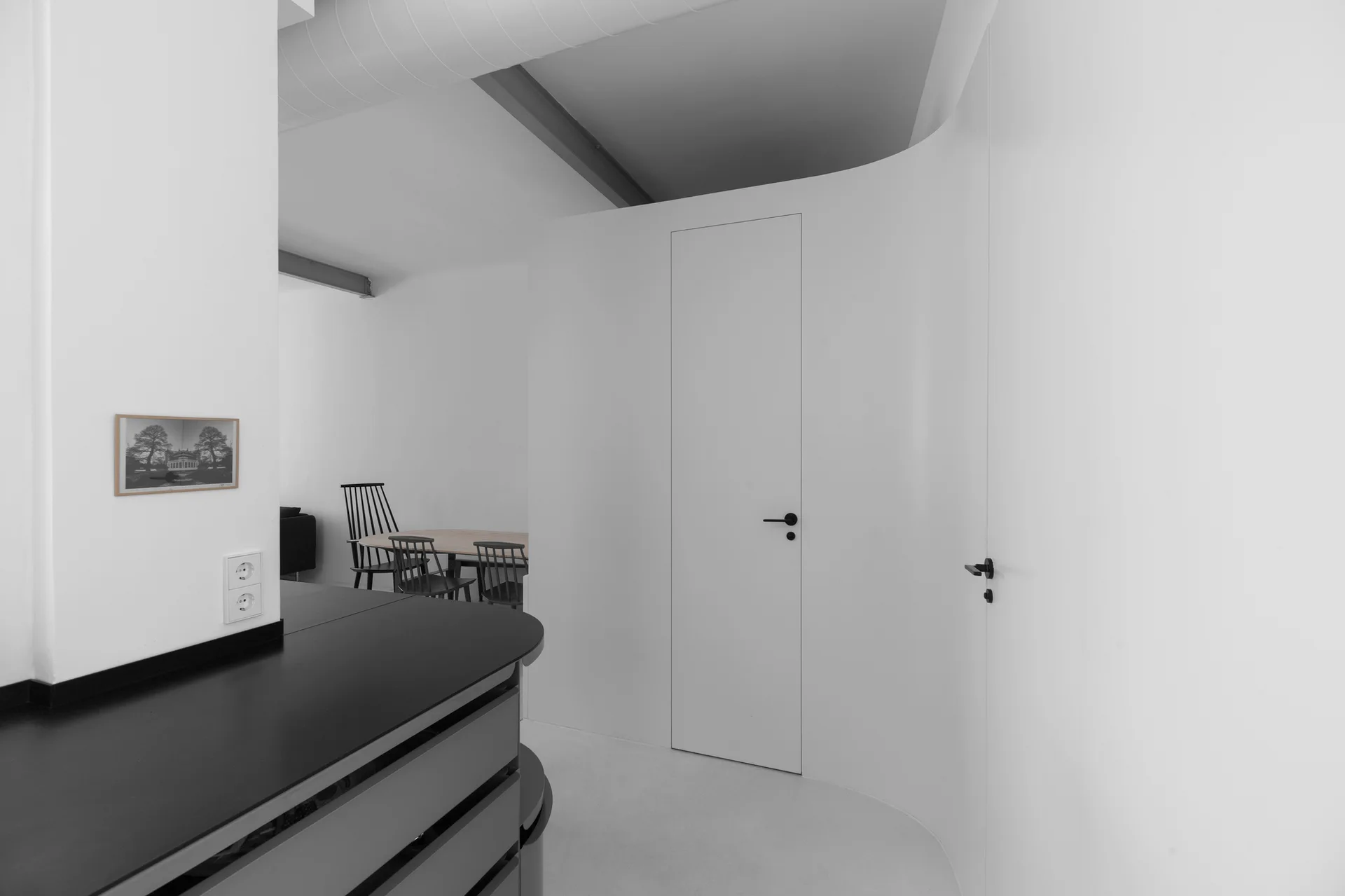A minimalist interior featuring a white curved partition wall with a flush door, a dark counter, and a background dining table with spindle chairs.