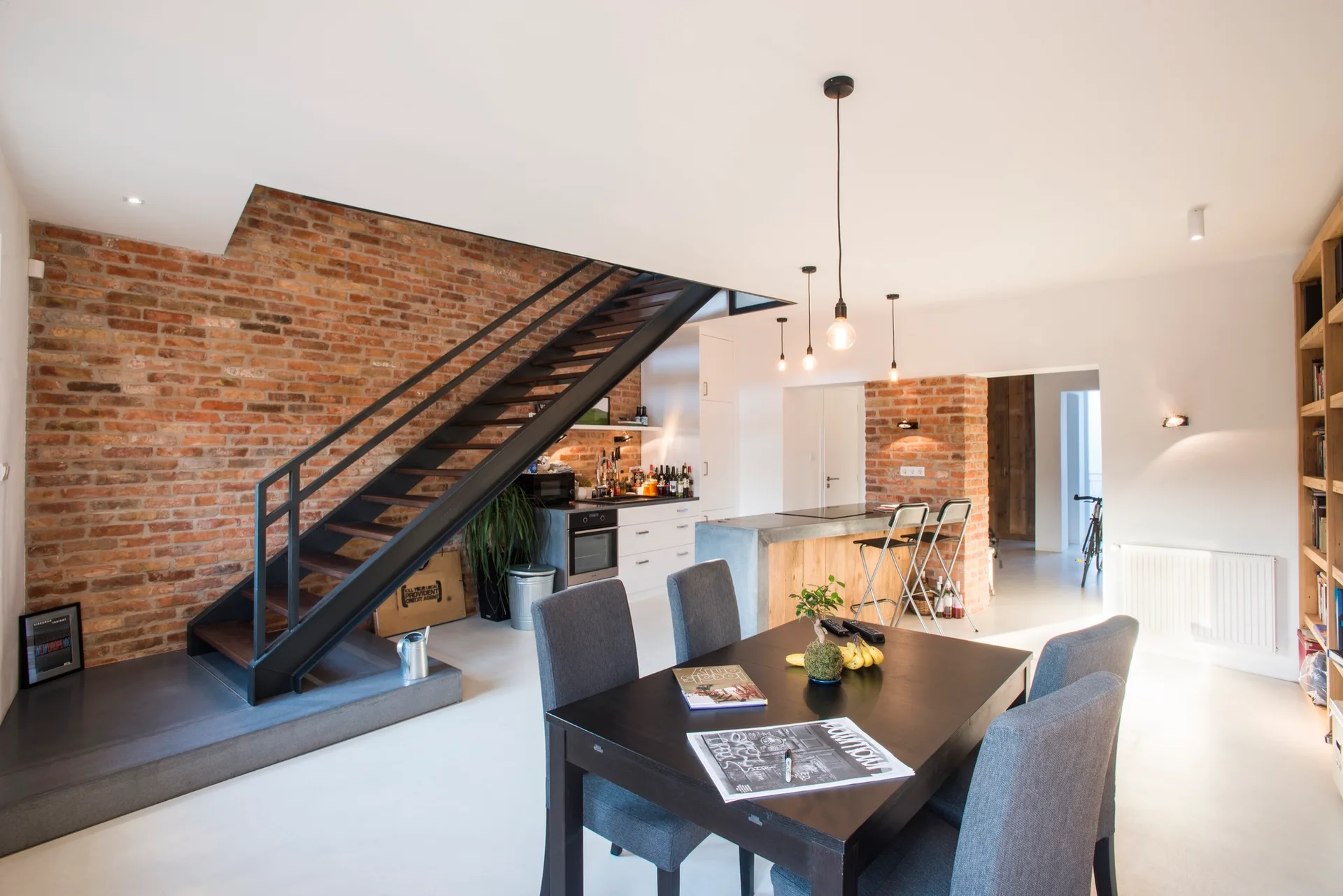 An open-concept dining area features a dark dining table with grey chairs, a black metal staircase, and an exposed brick accent wall.