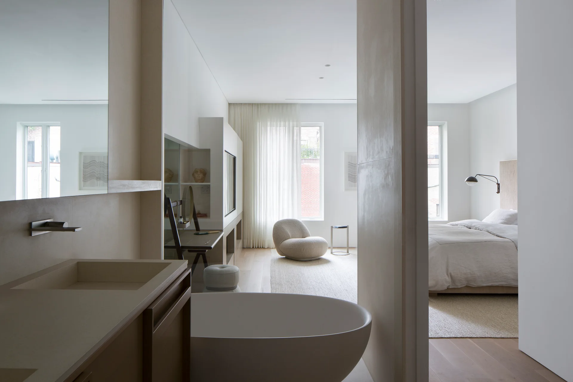 An open-concept suite shows a white freestanding bathtub and stone vanity in the foreground, with a bedroom and a curved beige armchair in the background.