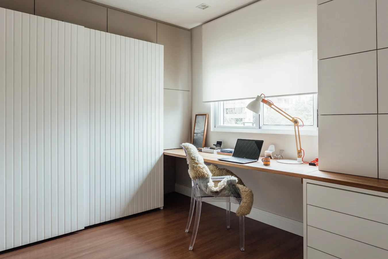 A minimalist home office features a long wooden built-in desk beneath a window, a clear acrylic chair with a sheepskin throw, and slatted white cabinets.