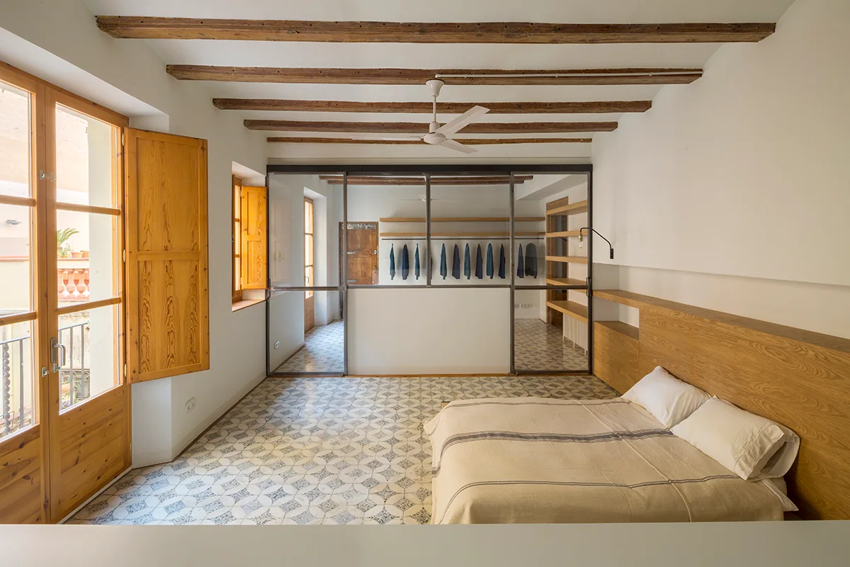 A rustic bedroom features a patterned tile floor, exposed wooden ceiling beams, and a slim glass partition separating the bed from a walk-in closet.