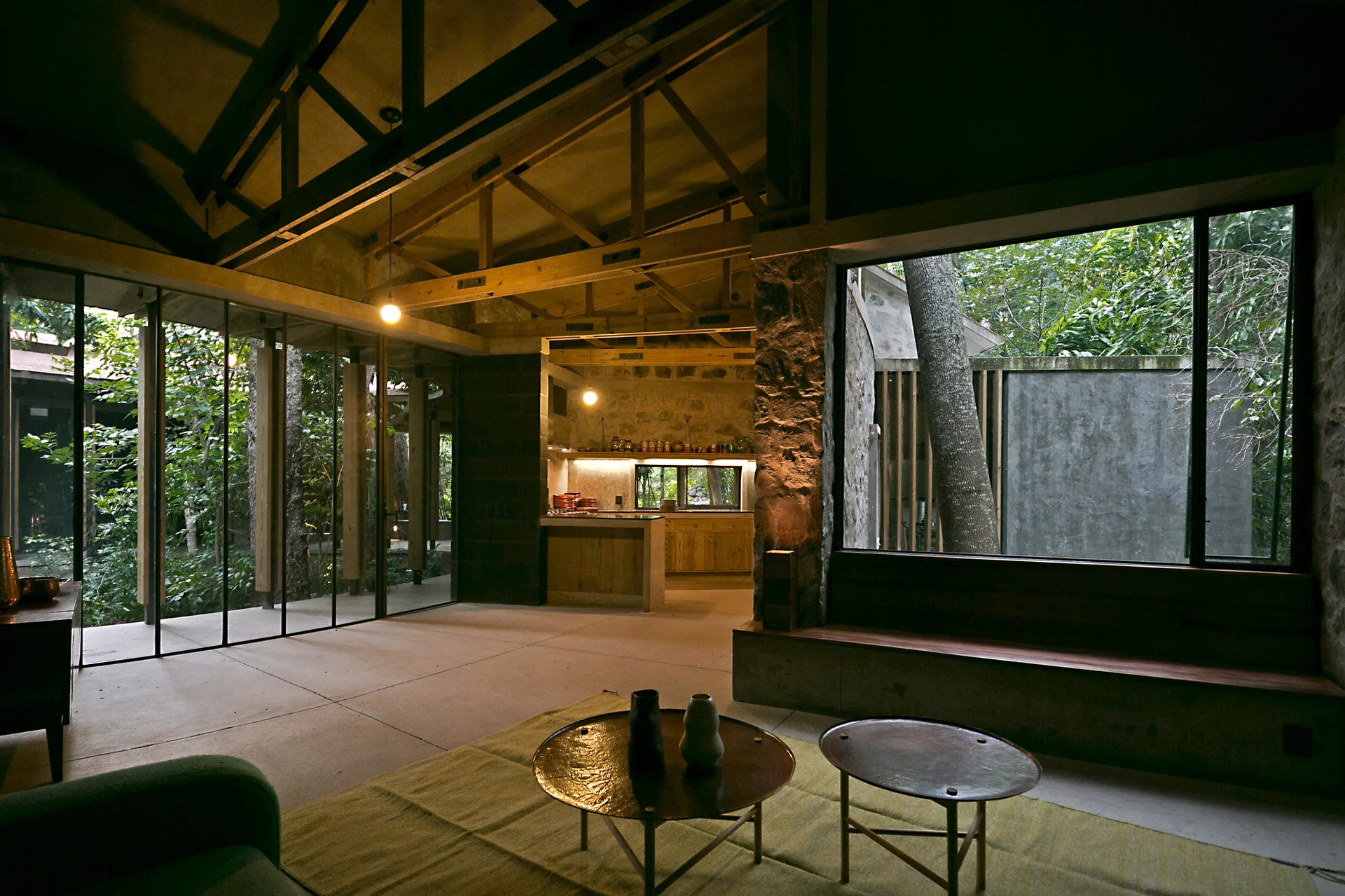 A rustic living room features exposed wooden ceiling trusses, textured stone walls, concrete floors, and large windows looking out onto a green garden.