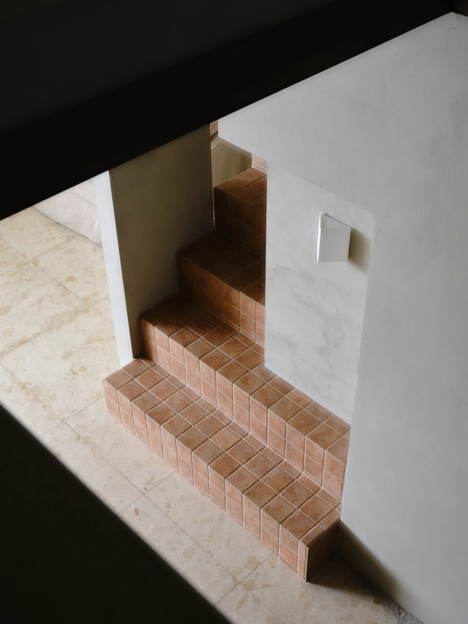 An overhead view of a staircase covered in square terracotta tiles next to smooth off-white plaster walls and a light stone tile floor.