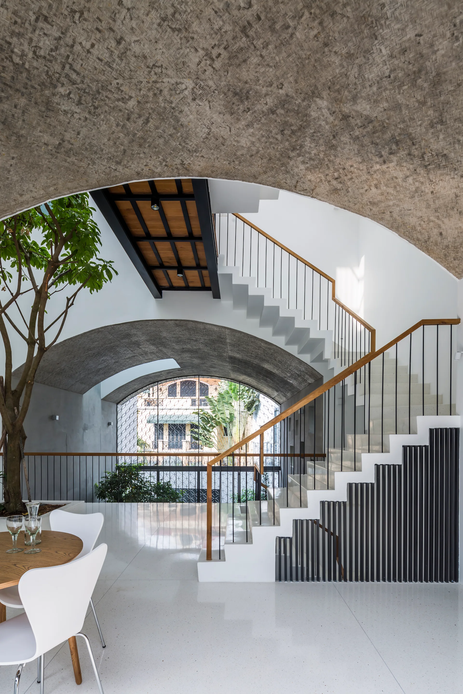 A white zigzag staircase with a wood handrail ascends beneath a textured concrete arched ceiling next to an indoor tree and polished white floors.