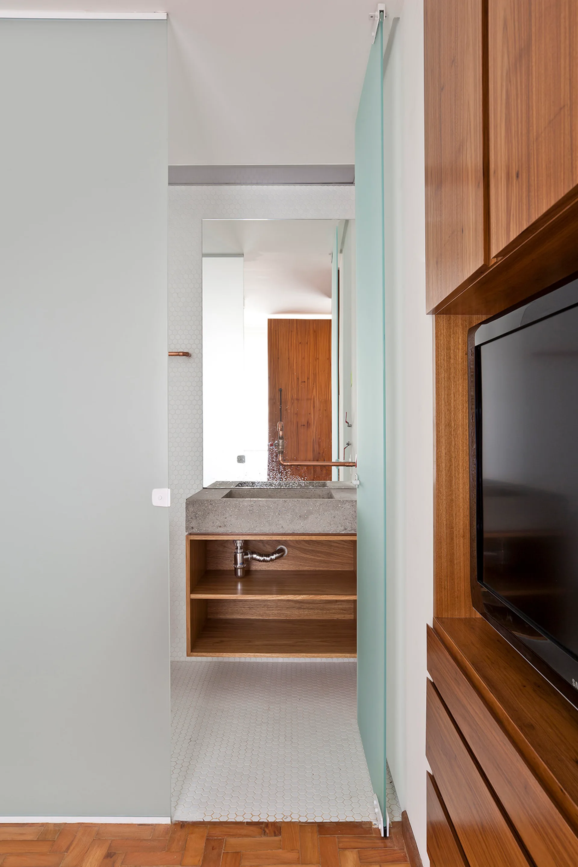 A frosted glass sliding door opens to a bathroom with a concrete basin sink, wood vanity, exposed copper pipes, and white hexagonal tiles.