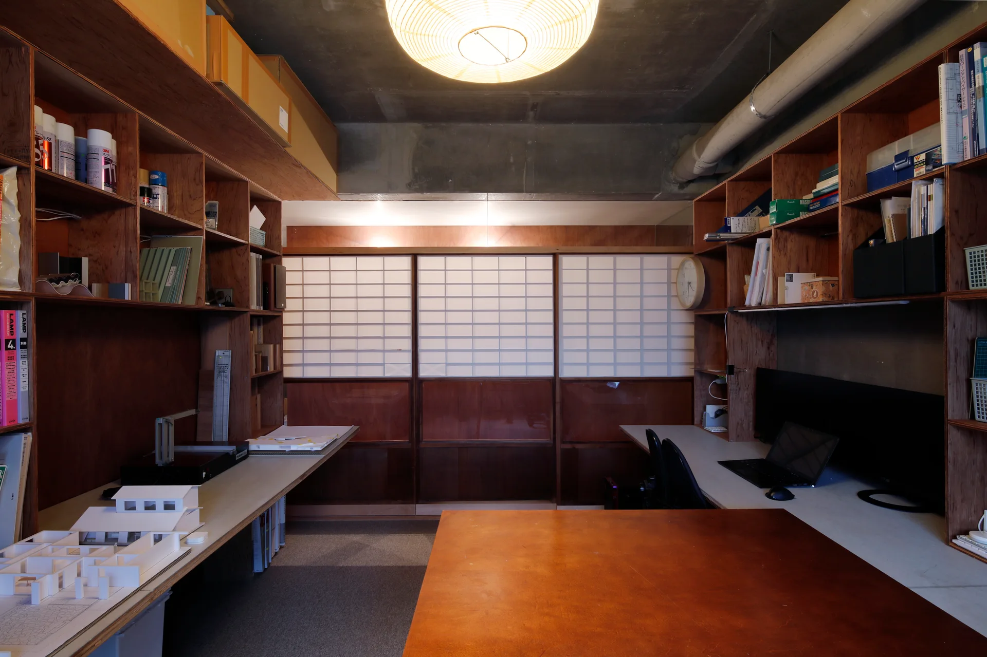 A home studio features built-in wooden shelves and desks, shoji sliding doors, and an exposed concrete ceiling with a round pendant light.