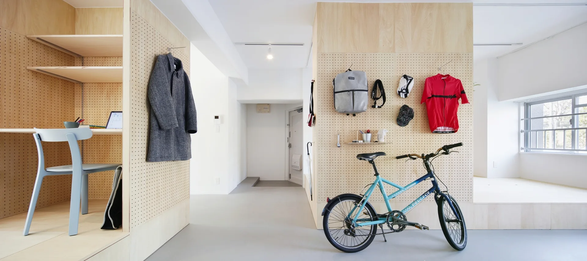 A bright entryway features light wood pegboard walls holding a coat, bag, and bicycle gear next to a built-in study nook with a blue chair.
