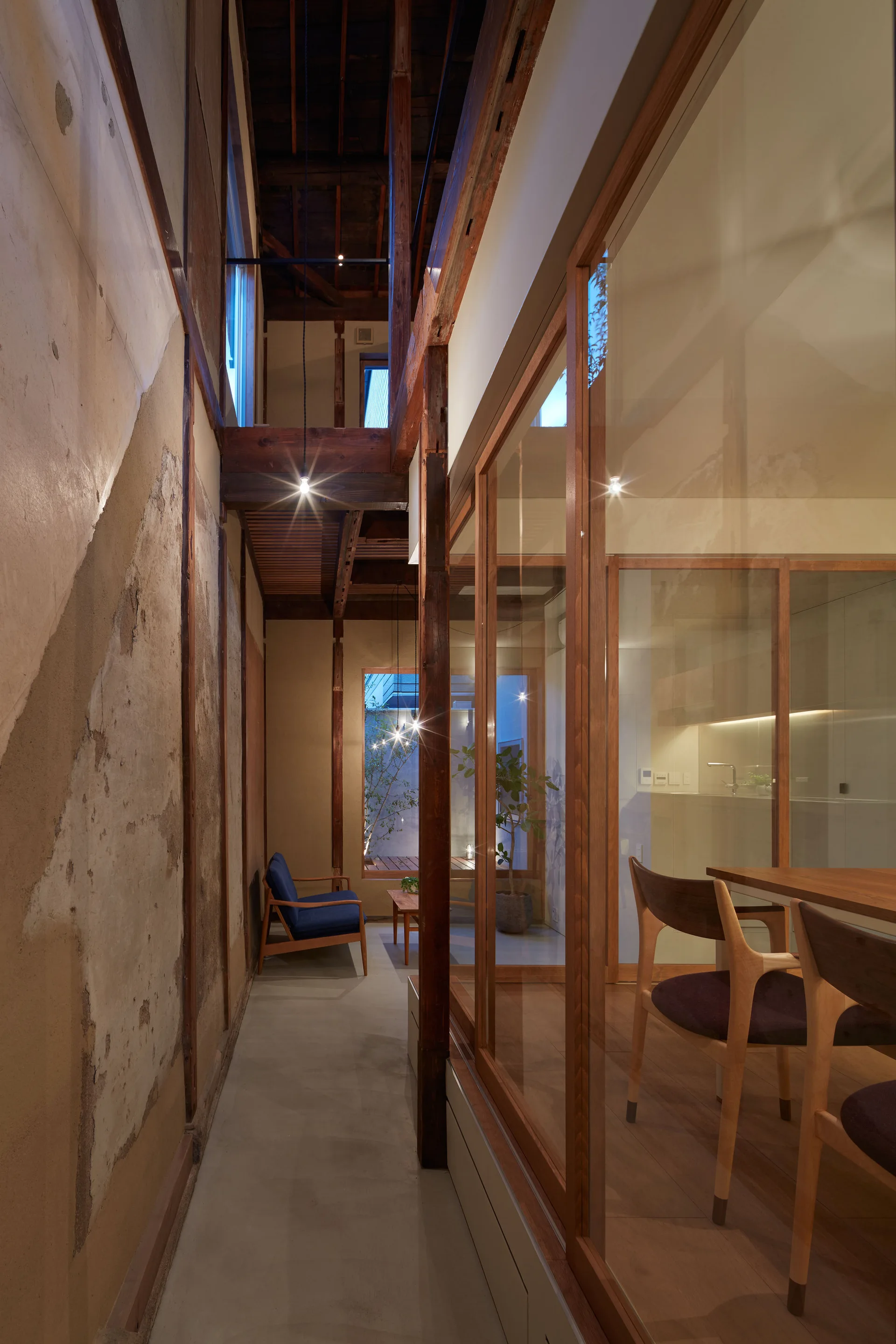 A narrow corridor with a distressed plaster wall and concrete floor runs alongside a glass-enclosed dining area with wood structural beams.