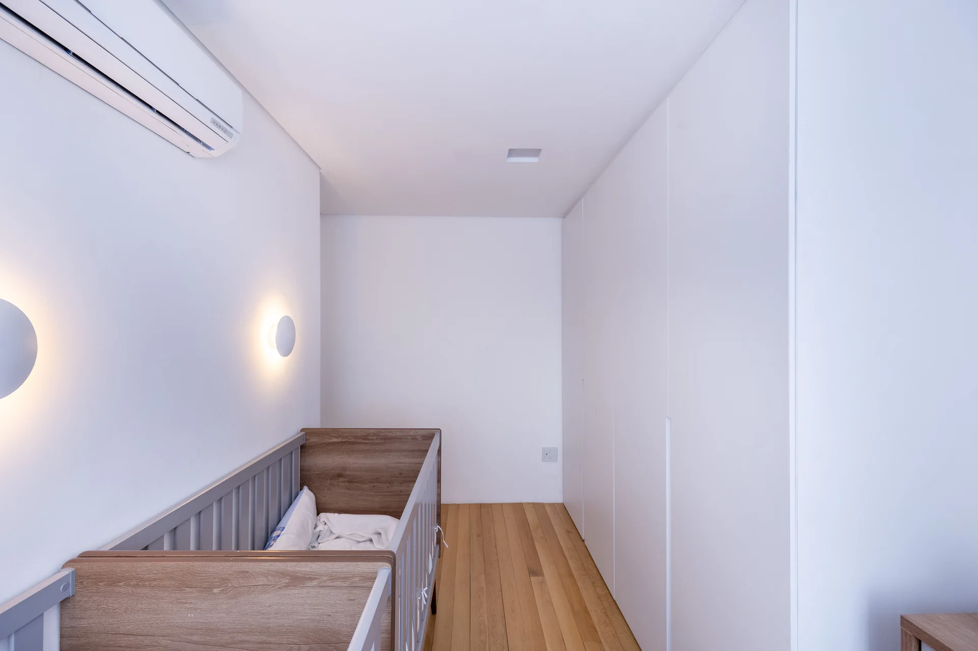 A minimalist white nursery featuring a wood-toned baby crib, circular wall sconces, wooden floors, and integrated floor-to-ceiling cabinetry.