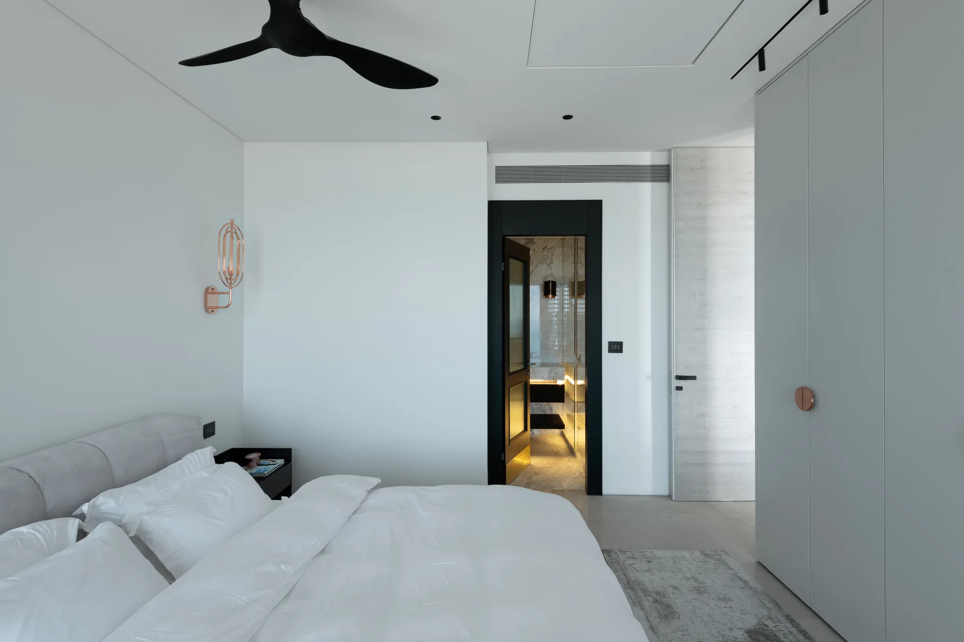 A minimalist bedroom features white bedding, a black ceiling fan, a copper wall sconce, and an open doorway showing a marble en suite bathroom.