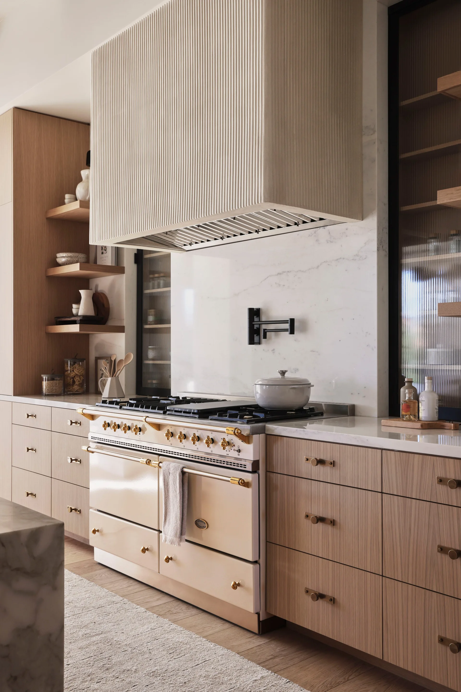 A cream-colored oven range stands below a textured fluted hood, bordered by light wood cabinets and a white marble backsplash.