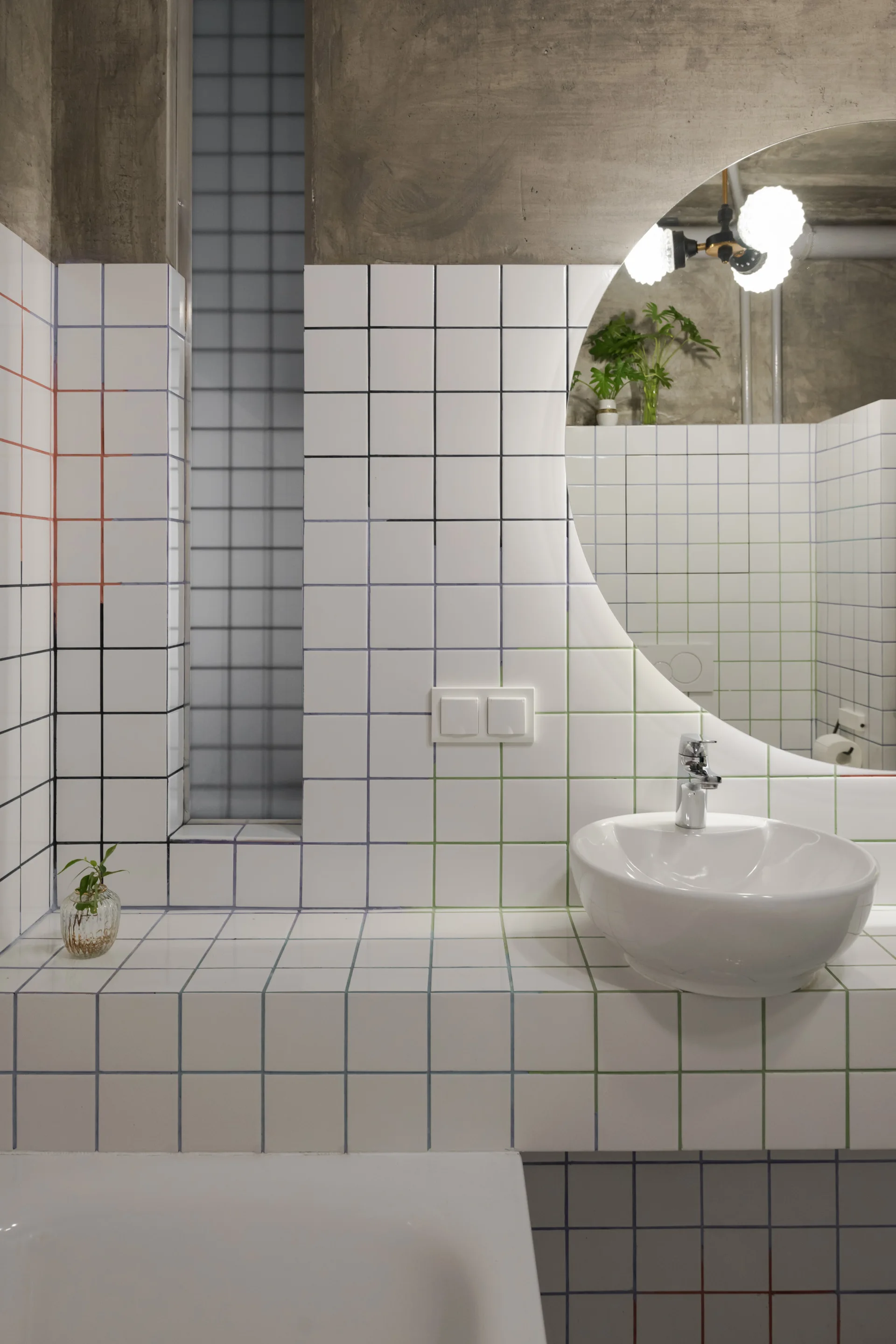 A bathroom vanity featuring a white vessel sink on a tiled counter with multicolored grout lines, a round mirror, and a concrete ceiling.