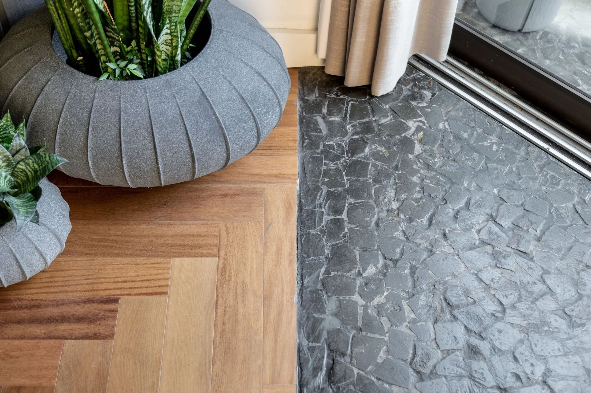 Warm herringbone wood flooring meets a dark, textured stone tile transition area featuring large gray fluted planters with green snake plants.