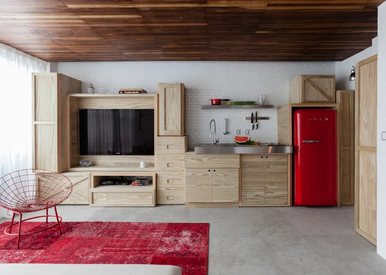 A combined kitchen and media area features light pine cabinets, a bright red refrigerator, a dark wood ceiling, and white subway tile walls.