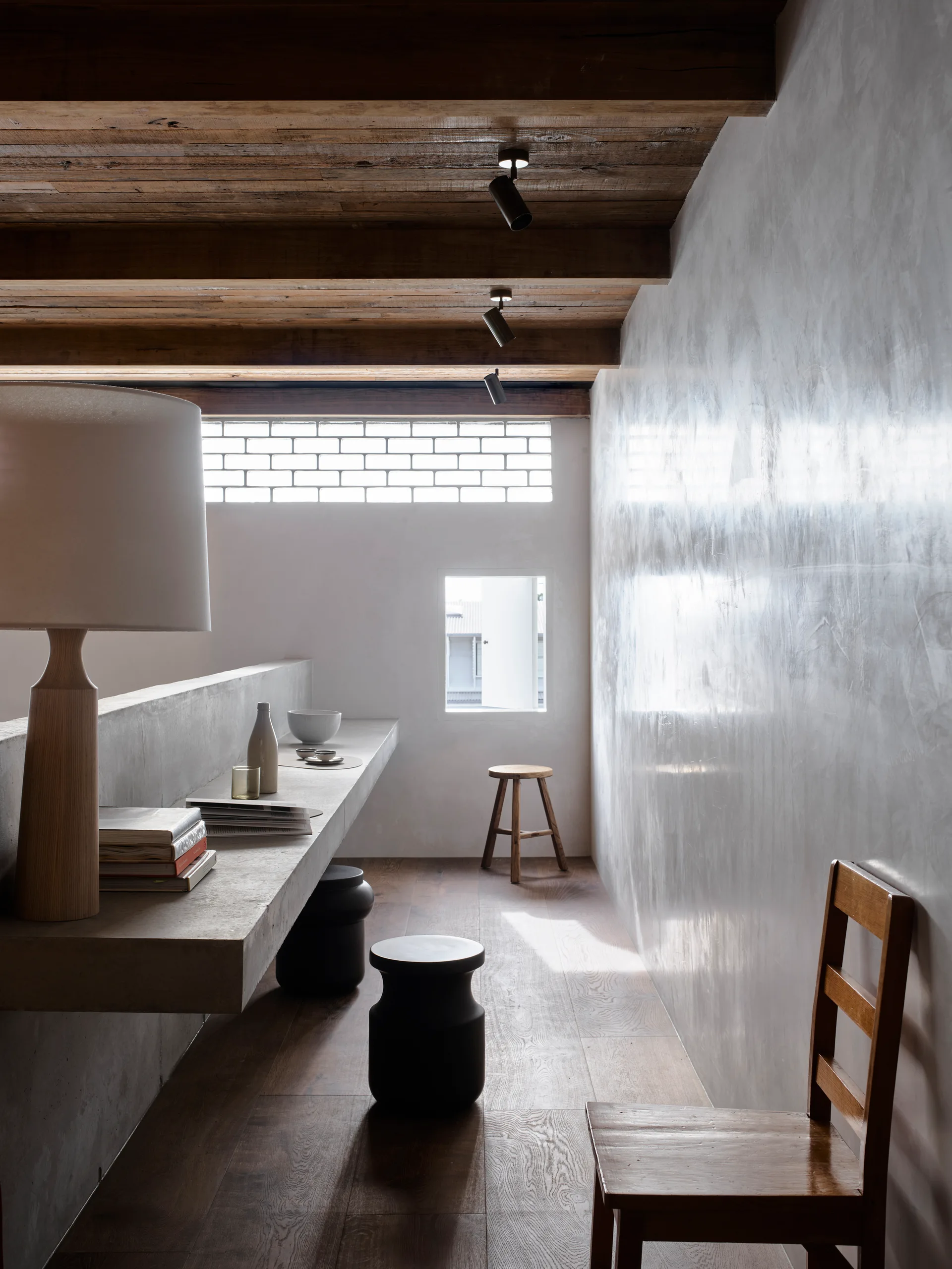 A minimalist loft corridor features a cantilevered concrete desk, a polished plaster wall, and exposed wooden ceiling beams with spotlights.