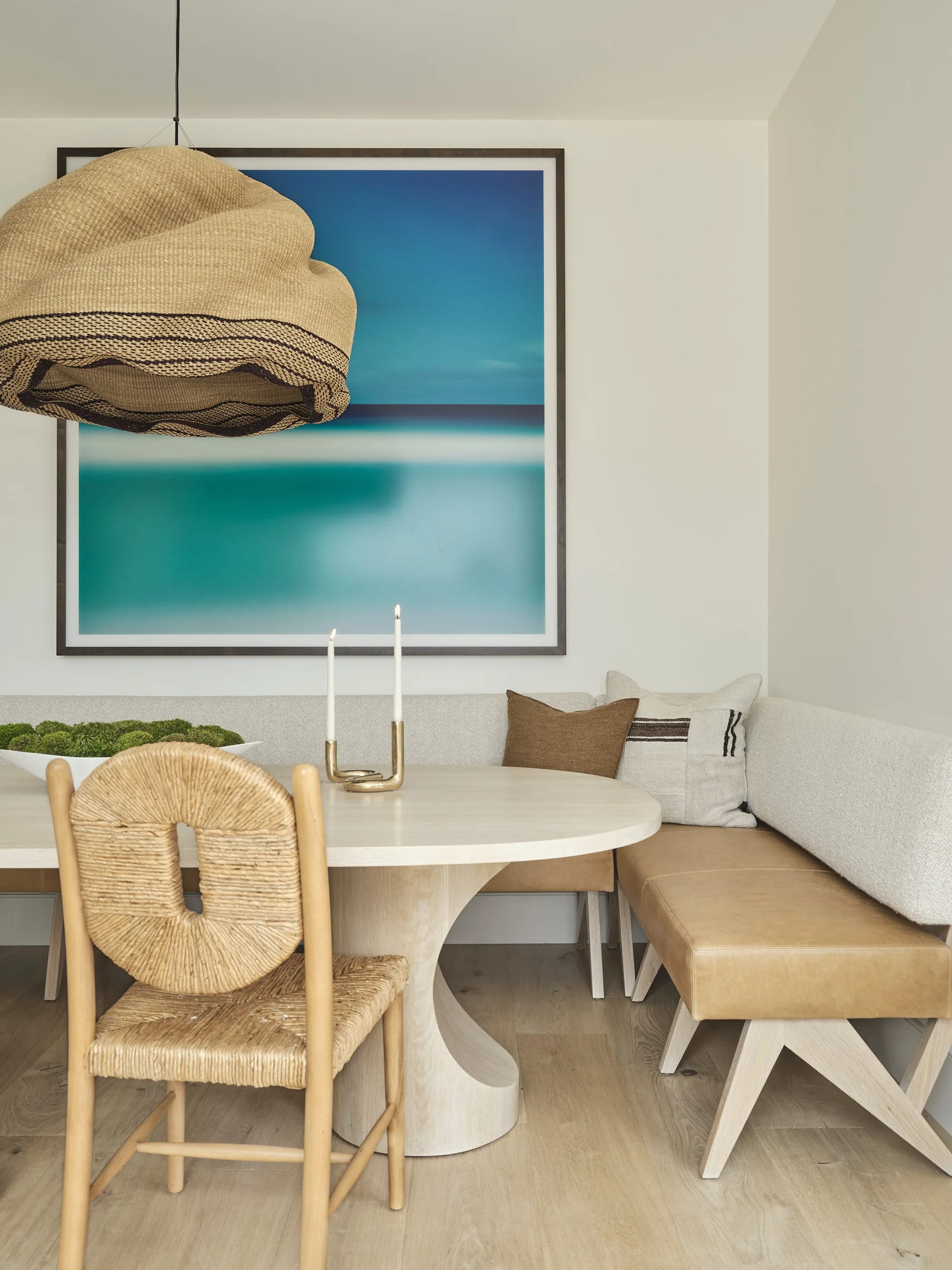 A modern breakfast nook features a light wood pedestal table, a tan leather corner banquette, a large woven pendant light, and a blue framed print.