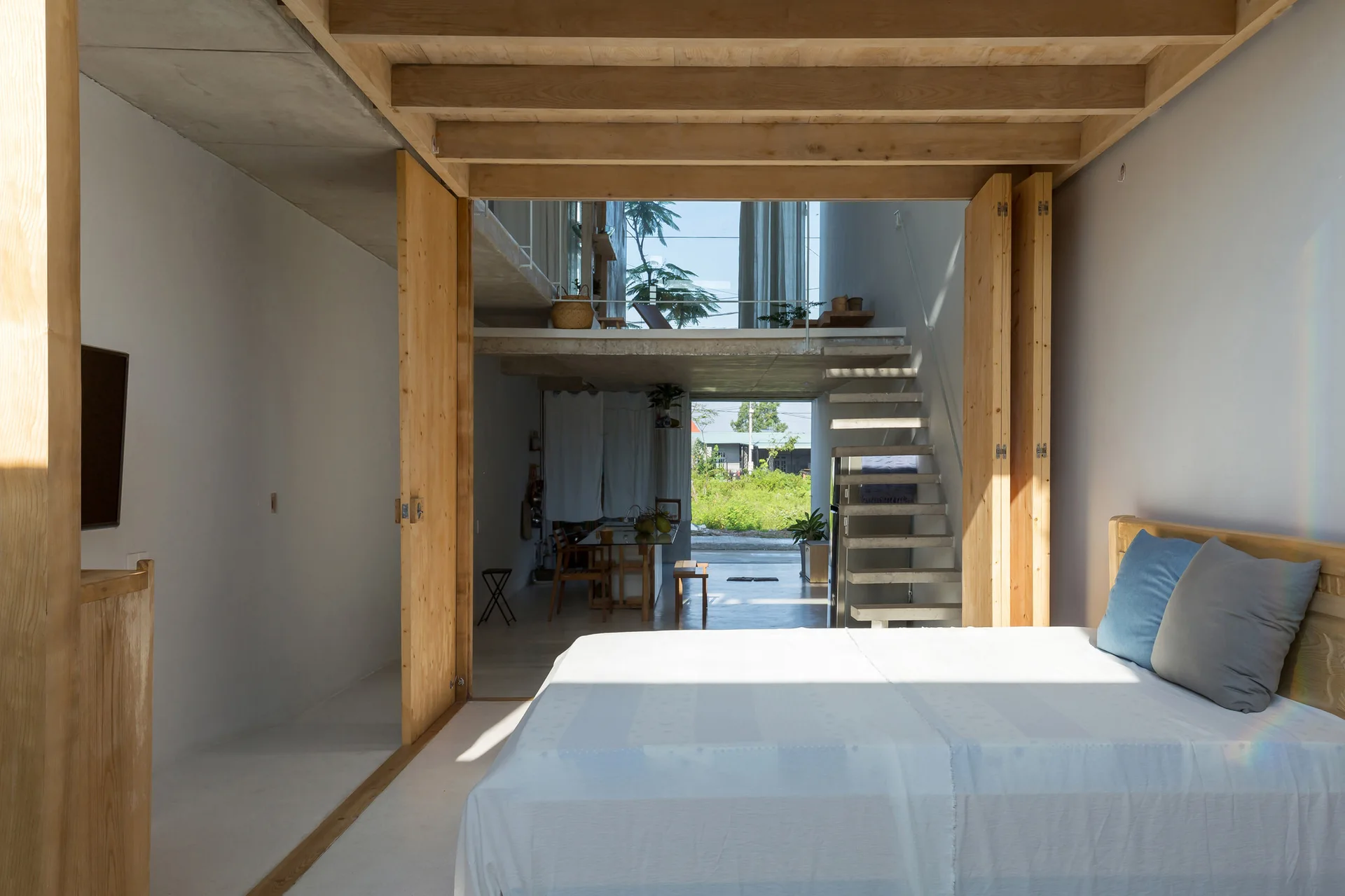 A minimalist bedroom with a white bed and blue pillows looks out through large wooden doors toward a concrete floating staircase in an open-plan home.