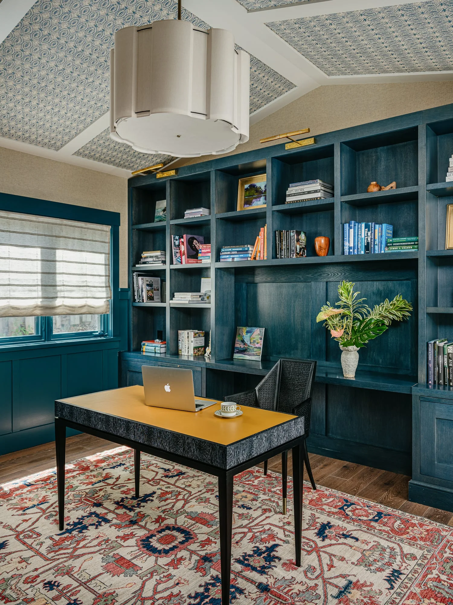 A home office features deep teal built-in bookshelves, a yellow-topped desk with a laptop, an ornate patterned rug, and wallpapered ceiling panels.