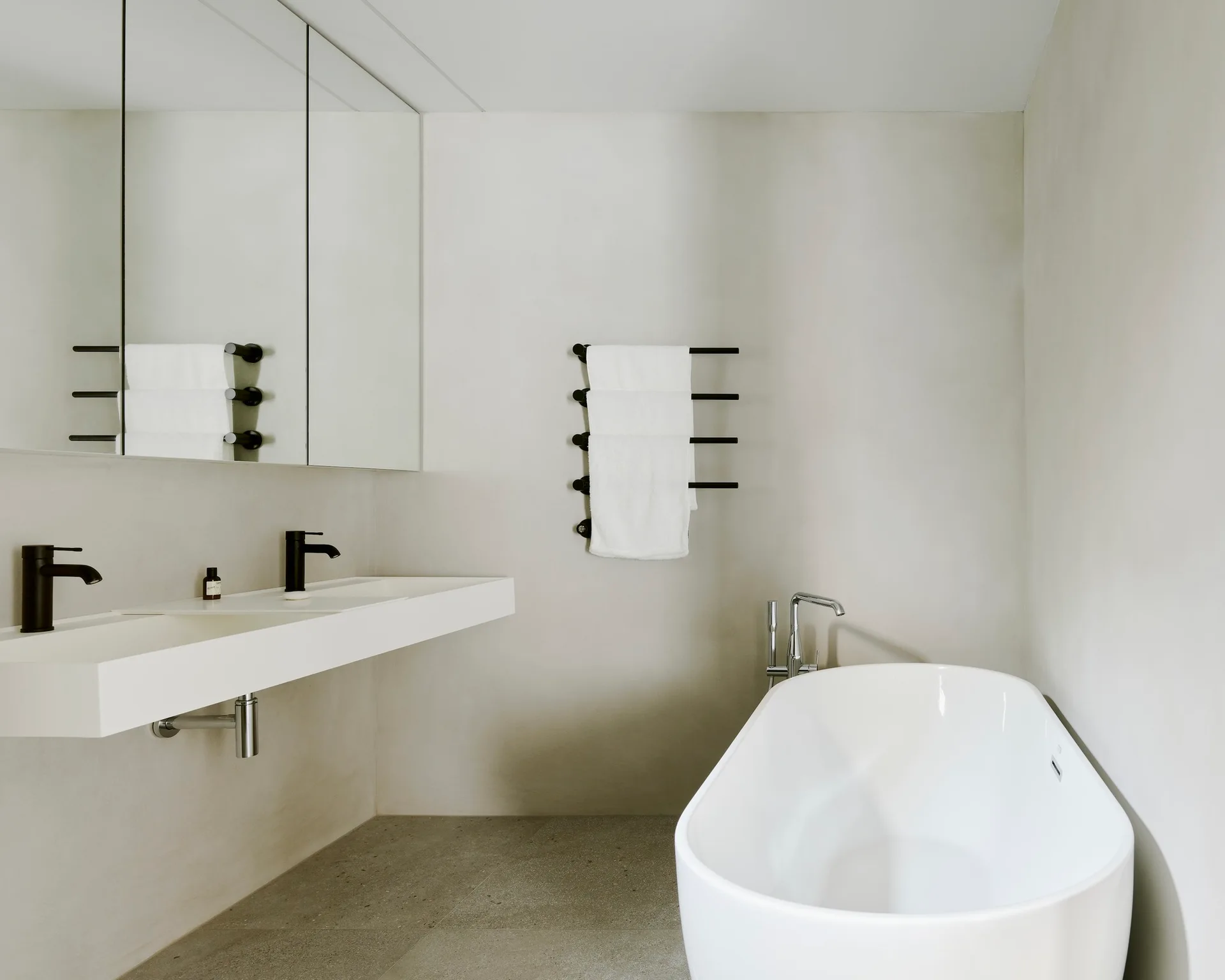 A minimalist bathroom with smooth light grey walls, featuring a white floating vanity, a freestanding oval bathtub, and a black ladder towel warmer.