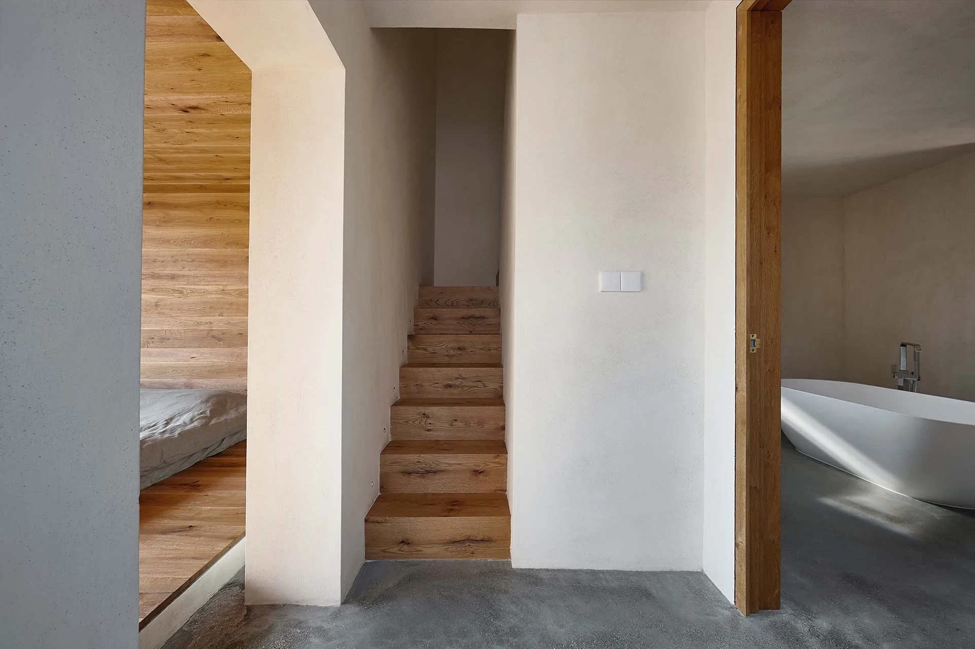 A minimalist hallway featuring a central wooden staircase, a bedroom with wood-clad walls on the left, and a bathroom with a white tub on the right.