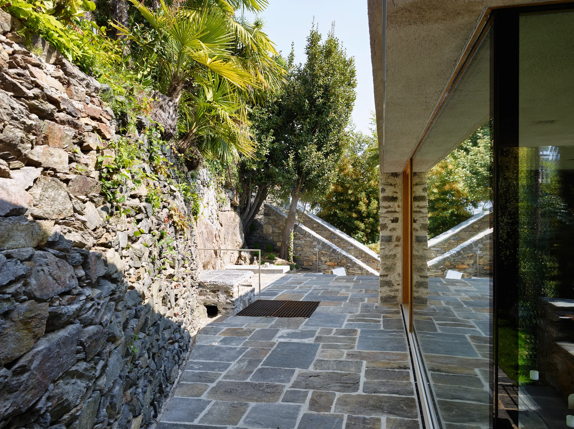 A stone-paved patio is bordered by a rugged natural stone retaining wall and a modern building with a large glass facade reflecting trees.