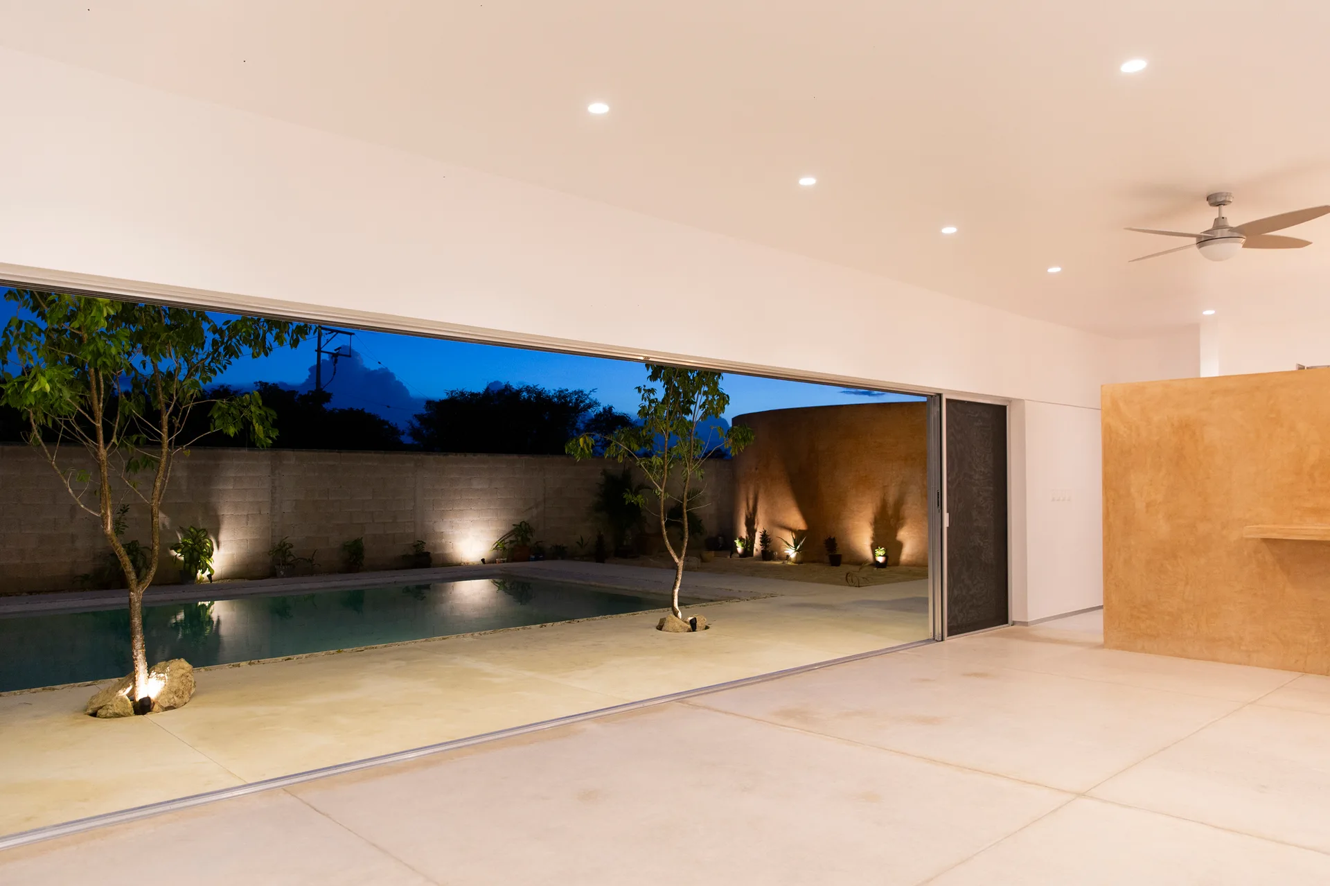 A wide sliding glass door opens a minimalist white room to a concrete patio with a swimming pool, small trees, and a curved tan wall at dusk.