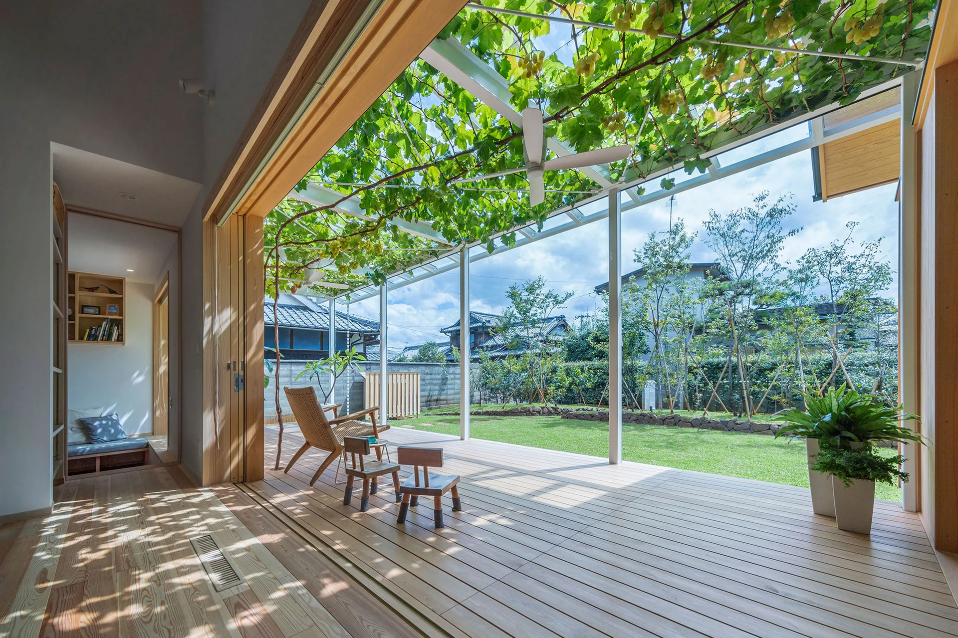 A light wood patio deck features a wicker armchair and small stools under a metal trellis woven with green grapevines next to a lawn.