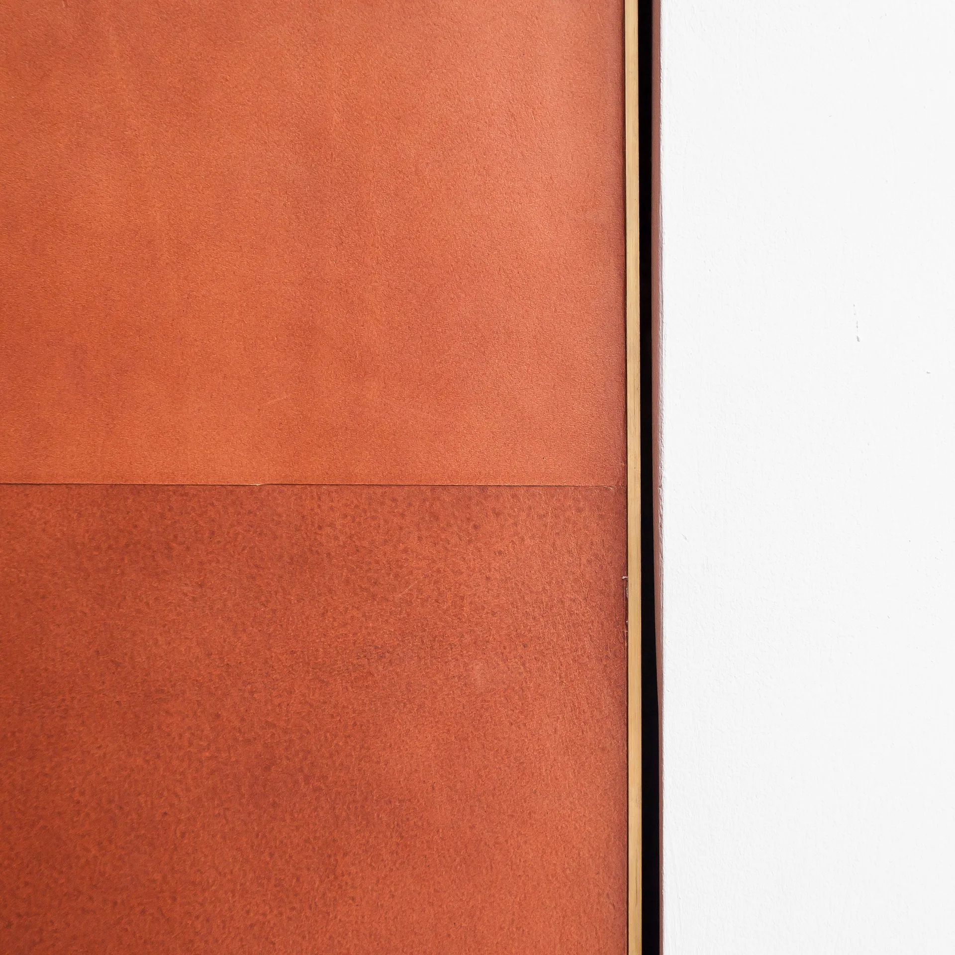 A close-up of terracotta leather wall panels featuring a subtle horizontal seam and a thin brass trim beside a smooth white wall.