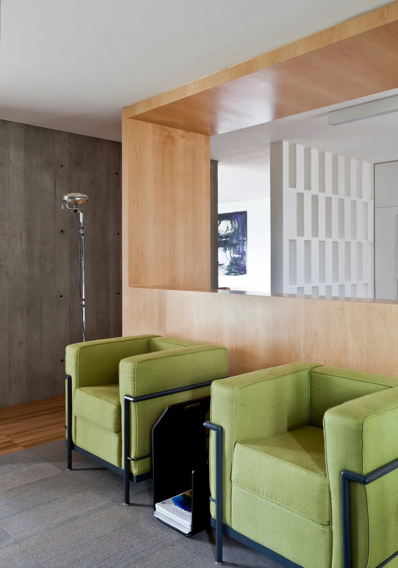 Two green upholstered armchairs with black metal frames stand on a gray rug next to a light wood room divider and a raw concrete wall.